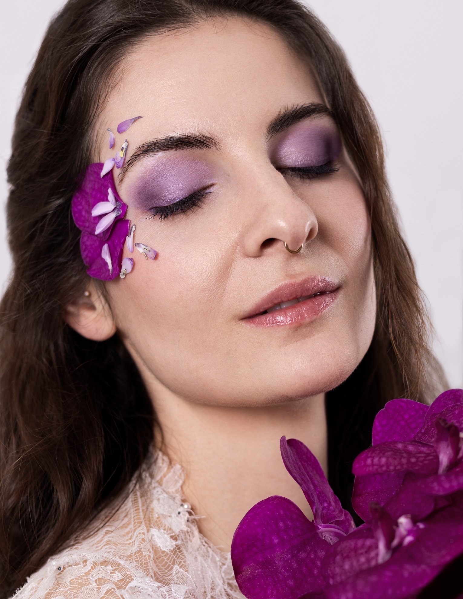 Nahaufnahme einer Frau mit lila Lidschatten, braunen Haaren, einem Nasenpiercing und einem weissen Spitzenkleid, umgeben von lila Blumen.