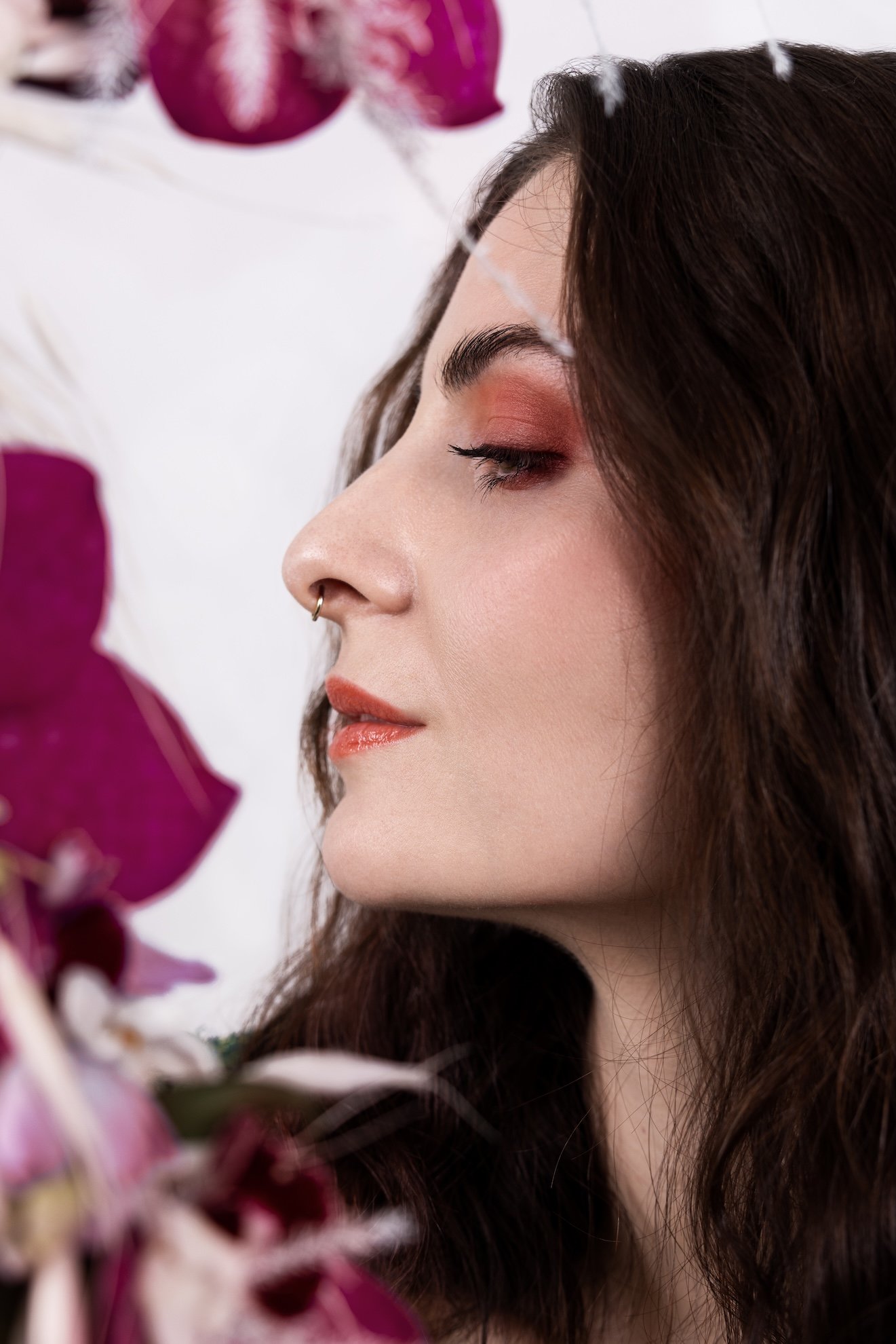 Eine Frau mit dunklen Haaren, geschminkt mit rosa Lidschatten und Lippenstift, die von Orchideen umgeben ist.