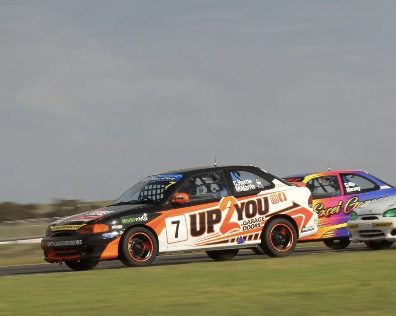 Race cars speeding on a track, with the black, orange, and white car labeled 'Up2You Garage Doors' in front, followed by colorful cars with sponsorship decals, under a cloudy sky.