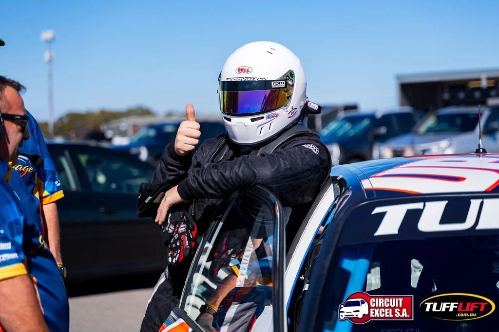 Race car driver in racing suit and helmet giving a thumbs-up while leaning on a race car at a race track.