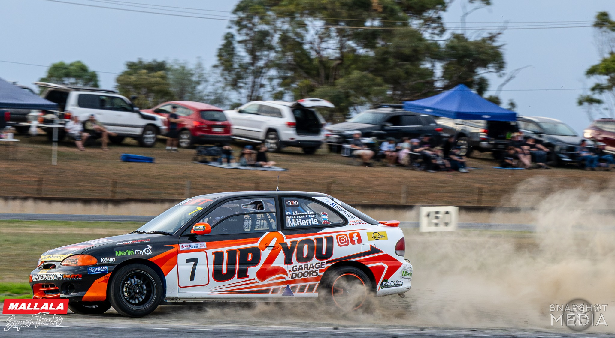 A race car speeding on a dirt track with spectators watching from the grassy hillside, some sitting under tents and canopies.