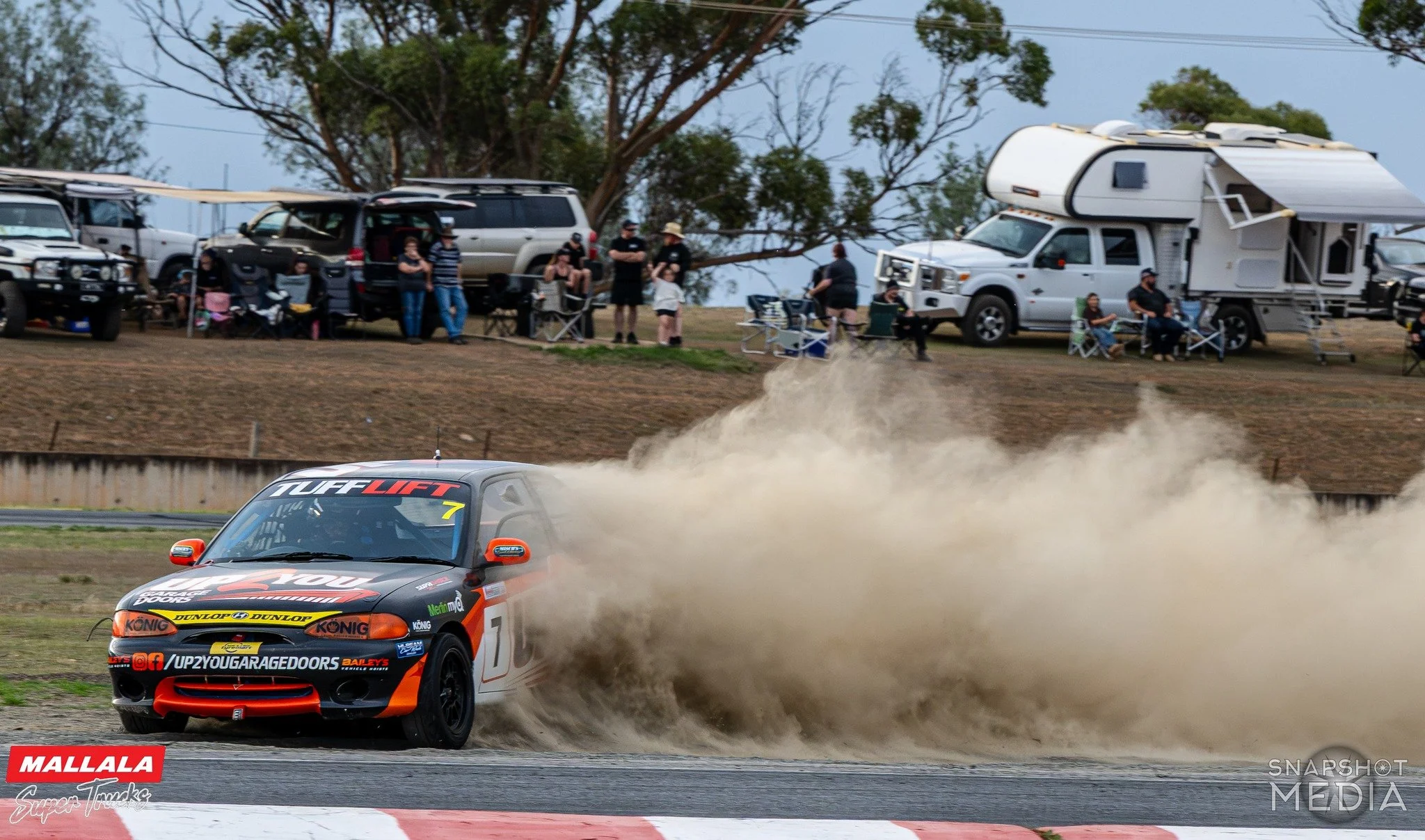 A race car kicking up dust on a dirt track during a motorsport event. Spectators are watching from the sidelines, some sitting in chairs and standing near parked vehicles. The scene is outdoors with trees in the background.