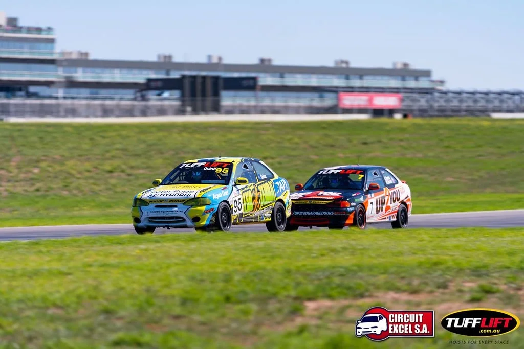 Two race cars on a track, one yellow and green with the number 95, and the other red and black with the number 100, competing in a race at a motorsport circuit.