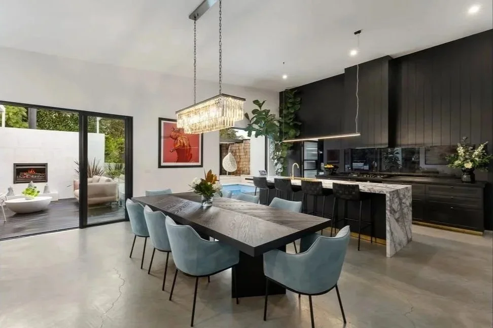Highett renovation kitchen and dining area with a long timber table, light blue chairs, black cabinetry and marble benchtops, opening onto an outdoor patio with a fireplace