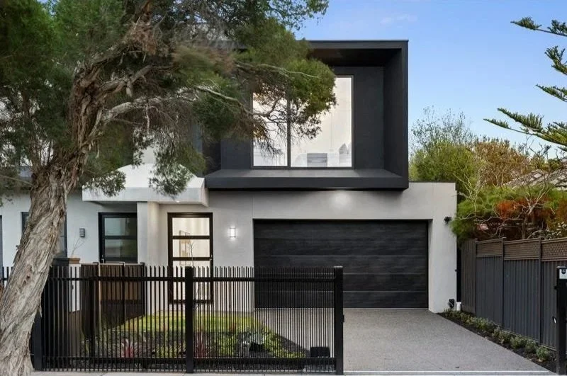 Modern two-story house with a white facade, black garage door, and large window box on the upper level. A small front yard with a black fence and gate, trees, and shrubbery surrounds the property.