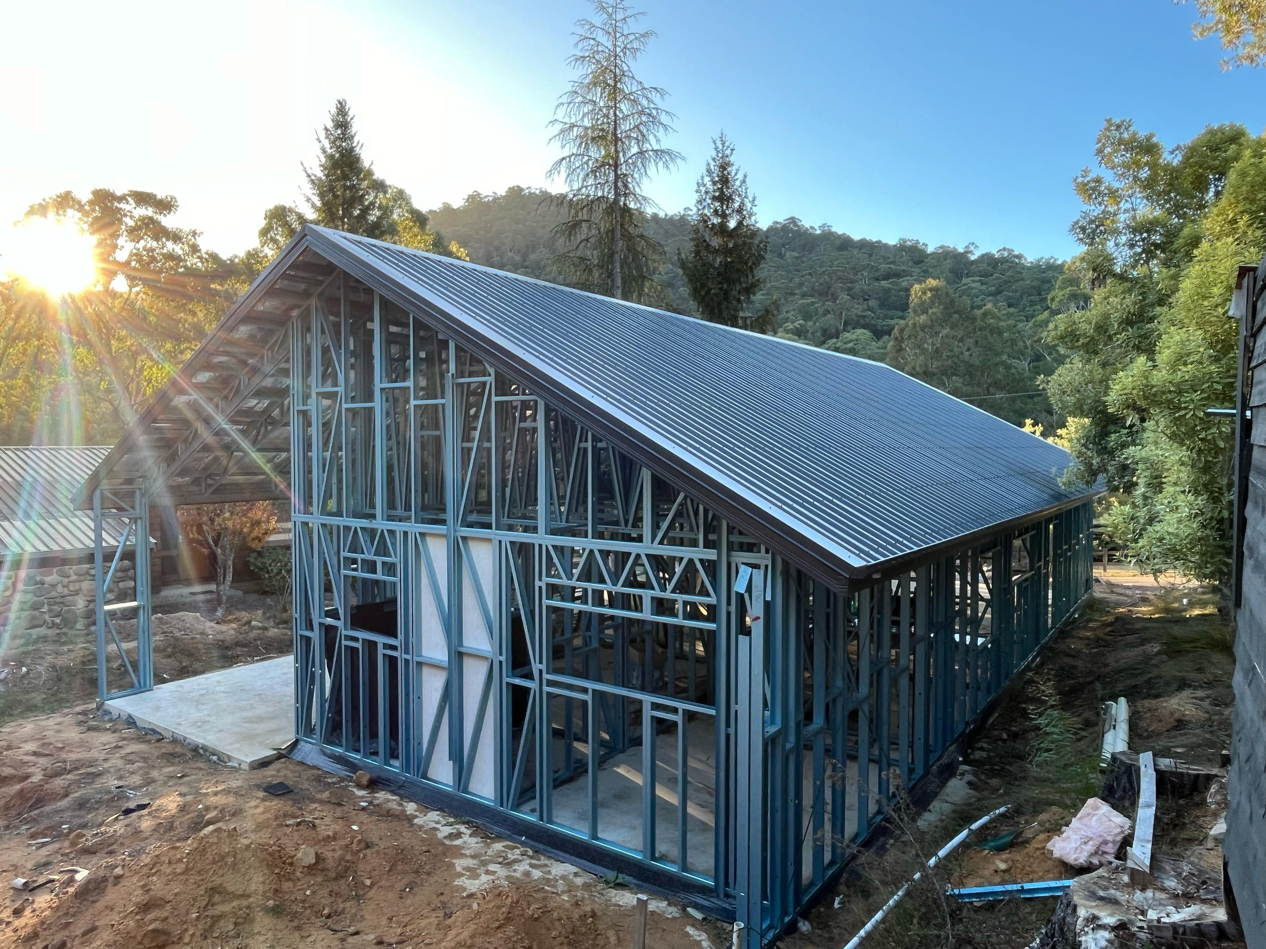 Alpine kit home under construction near Mansfield, with the steel frame and roof on, surrounded by trees and mountains in the late-afternoon sun