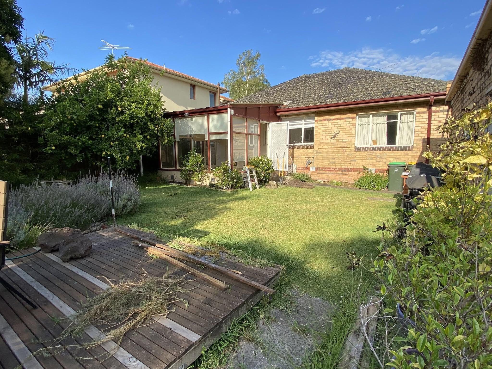 McKinnon backyard before the renovation, with the old timber deck lifted, a lawn and garden beds behind the brick home