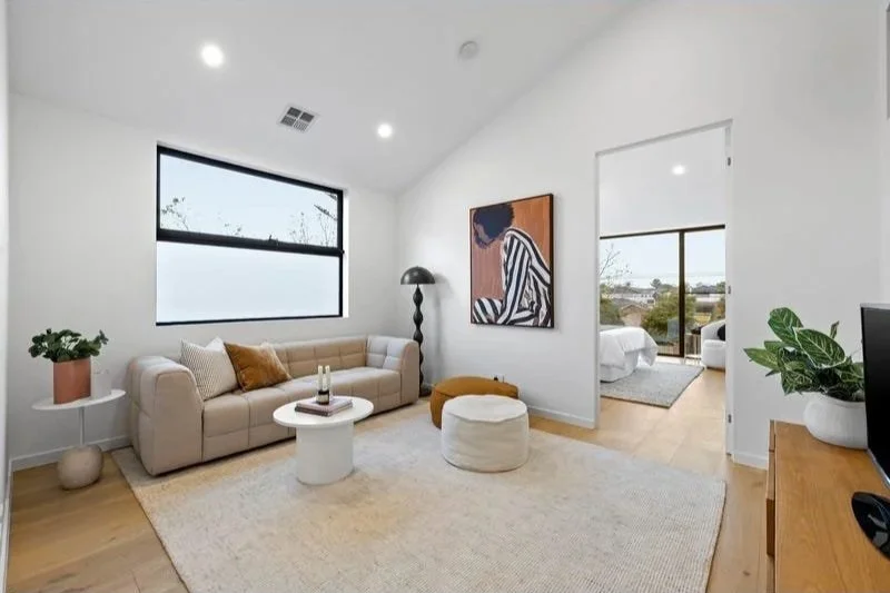 Modern living room with beige sofa, white coffee table, abstract artwork on white wall, potted plant, floor lamp, window with view, and a doorway leading to a bedroom.