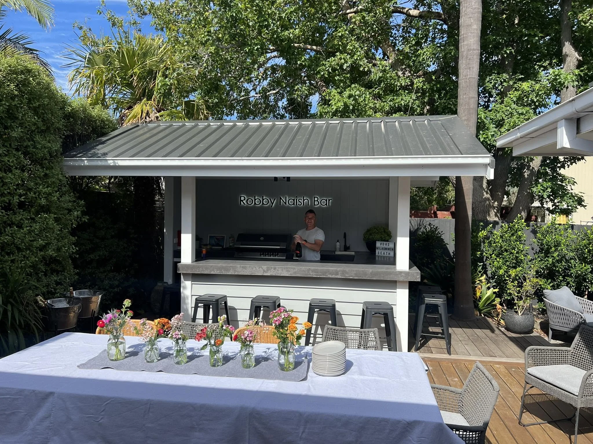 An outdoor gathering area with a long table covered with a white cloth, decorated with small vases of colorful flowers, surrounded by chairs, and a bar area in the background with a man standing behind it. Tall trees and lush green foliage surround the space.