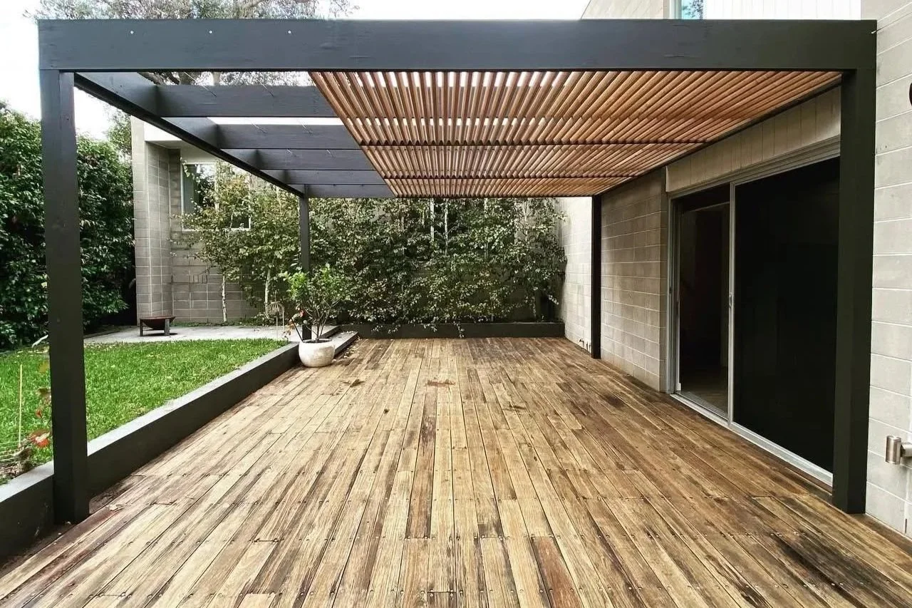 Wooden decked outdoor patio area with a black metal pergola and lattice ceiling, adjacent to a house with glass sliding doors. Green plants and trees in the background.