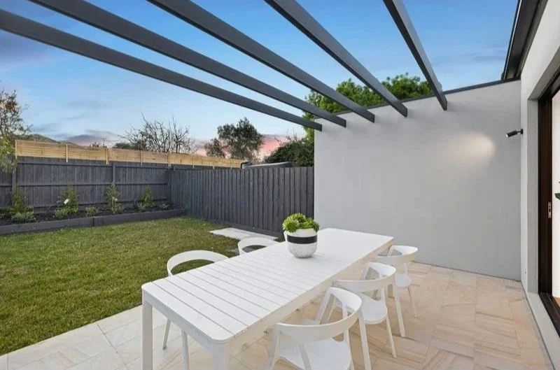 Modern outdoor patio with white dining table, six matching white chairs, a potted plant, and a glass sliding door leading to the house. The patio has a tiled floor and is partially covered with a pergola, overlooking a grassy yard with a wooden fence and small garden bed.