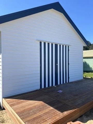 Rebuilt beach boatshed with white weatherboard cladding, a striped timber door and a new deck