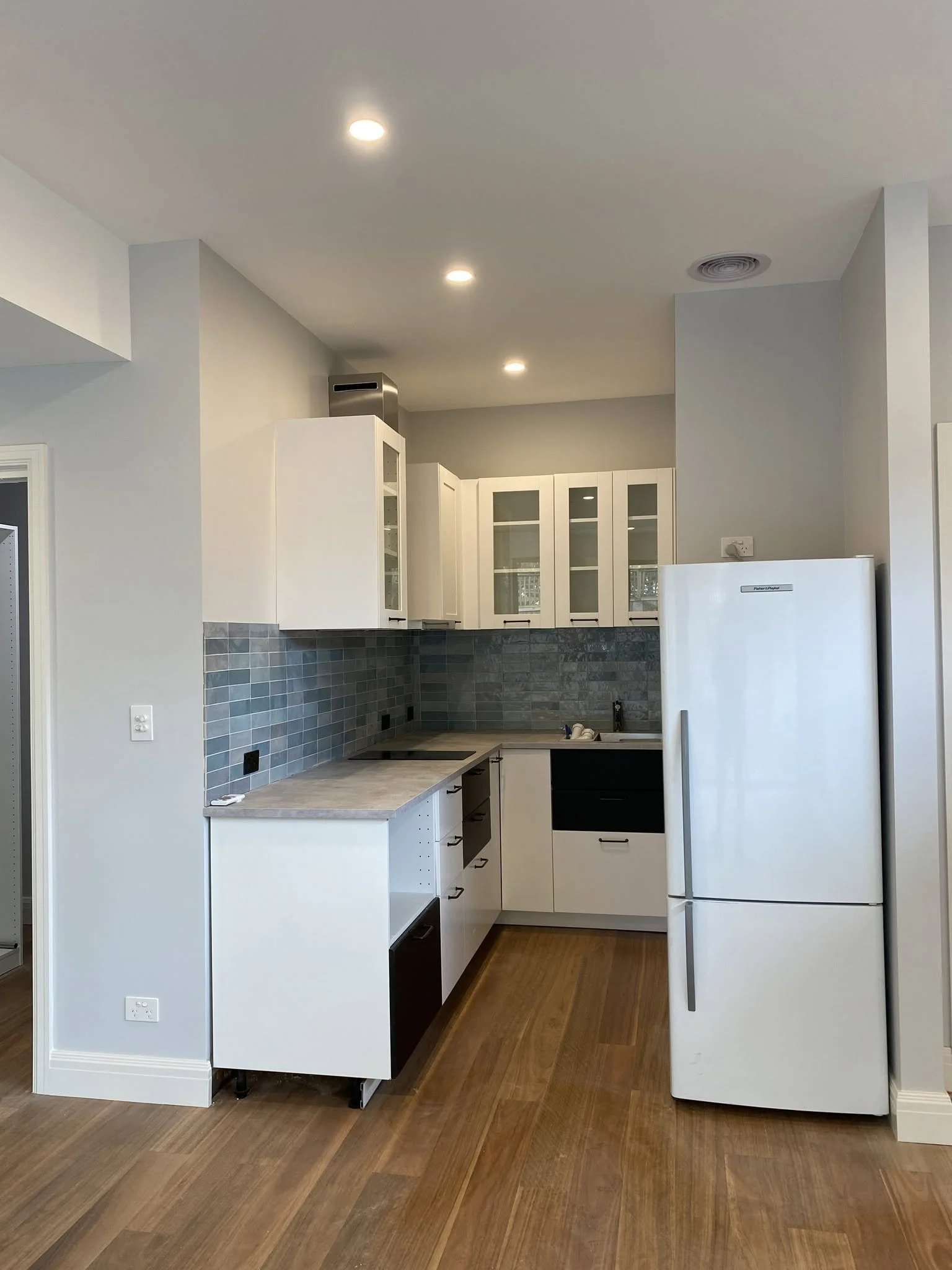 Kitchenette in the self-contained unit with white cabinets, a blue tiled splashback and a fridge