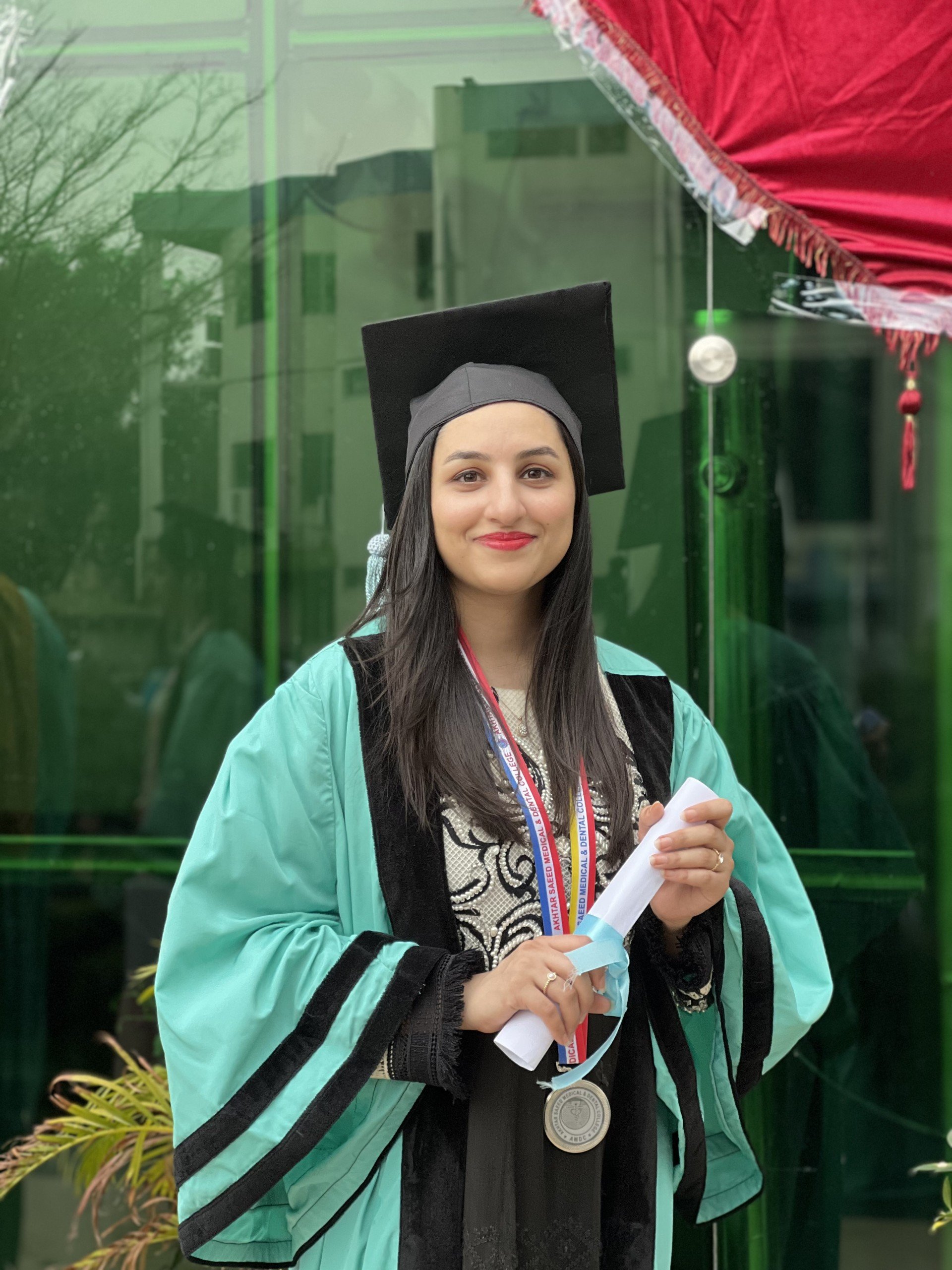 A young woman in a medical graduation gown and cap, holding a rolled diploma and wearing a medal, standing outdoors in front of a glass window with trees and buildings reflected in it.