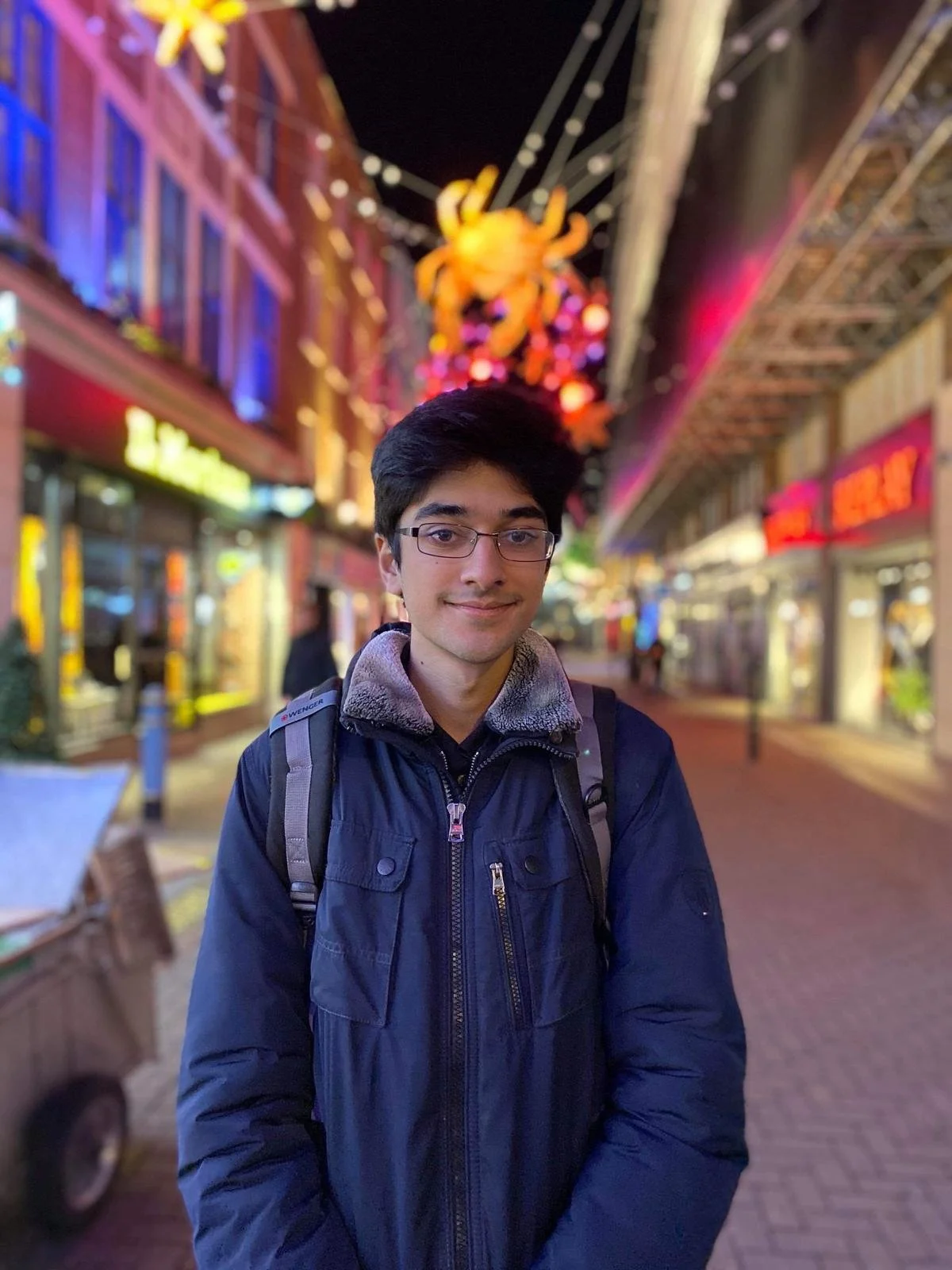 A young man with glasses and a jacket standing on a vibrant city street at night with lights and a large illuminated crab decoration overhead.