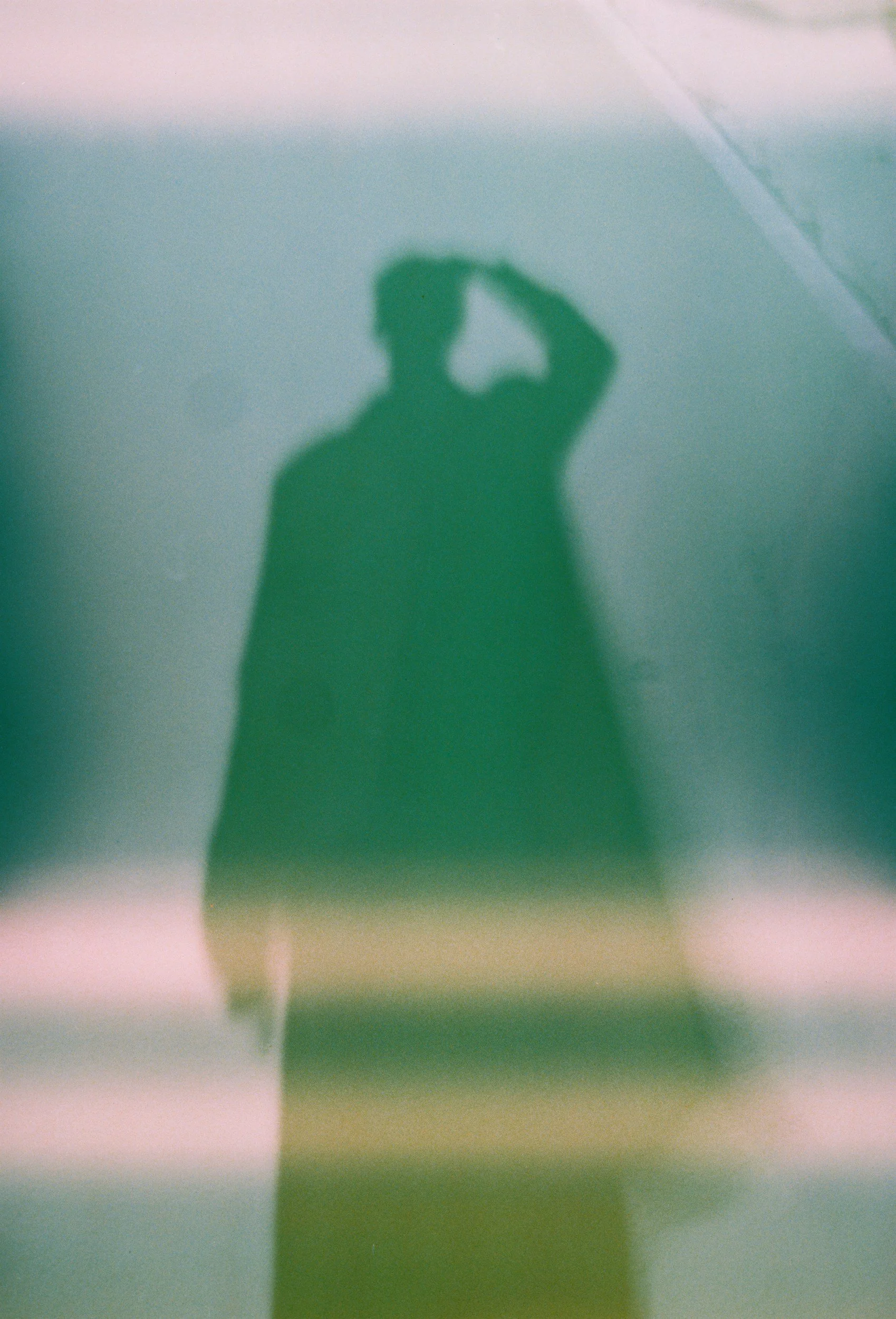A blurry shadow of a person taking a photo, with a slightly vertical figure and hand near their head, cast on a light green wall.