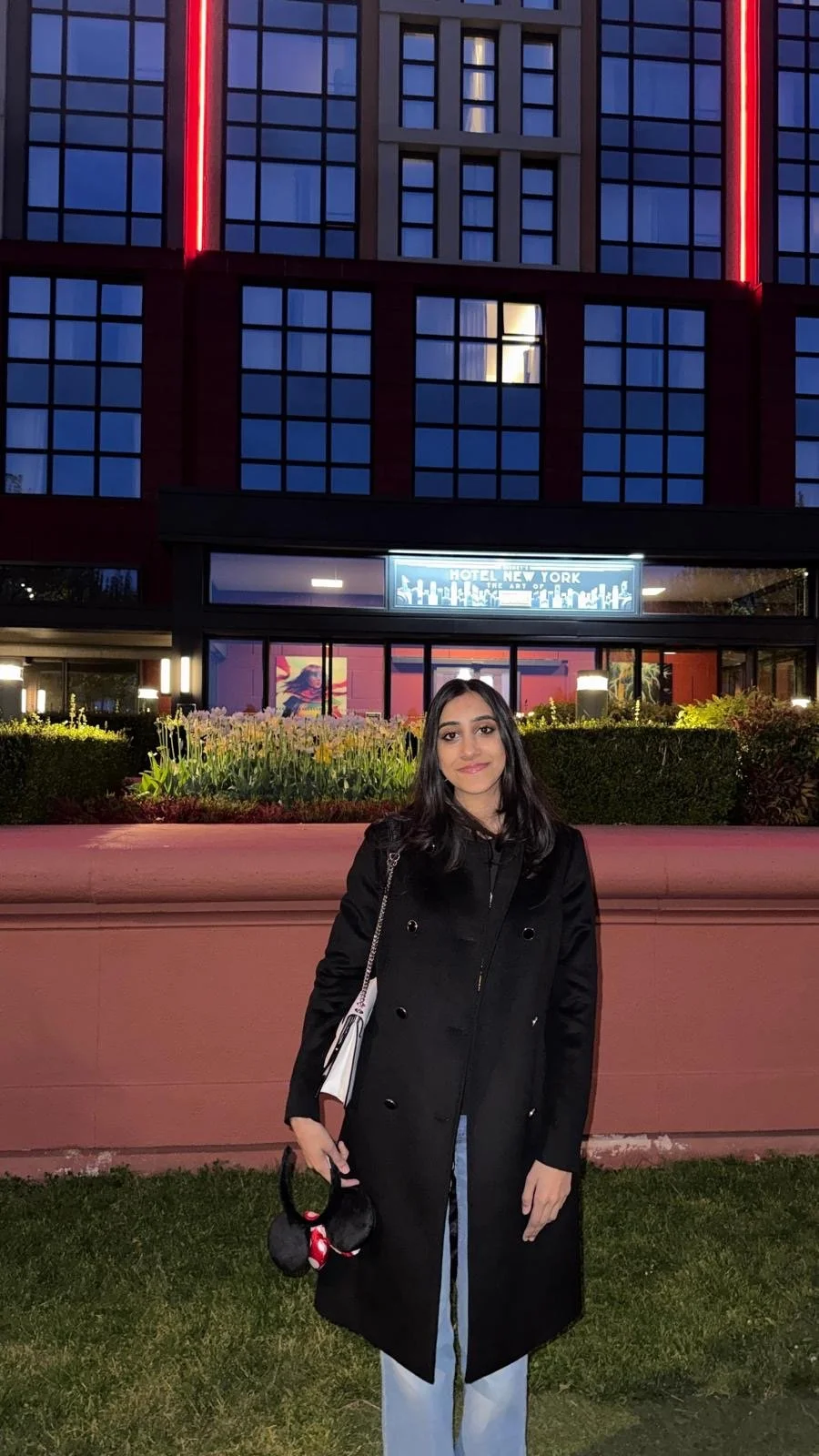 A young woman standing outside in front of a building with illuminated signs at dusk. She is wearing a black coat and holding a white bag and a pair of black headphones.