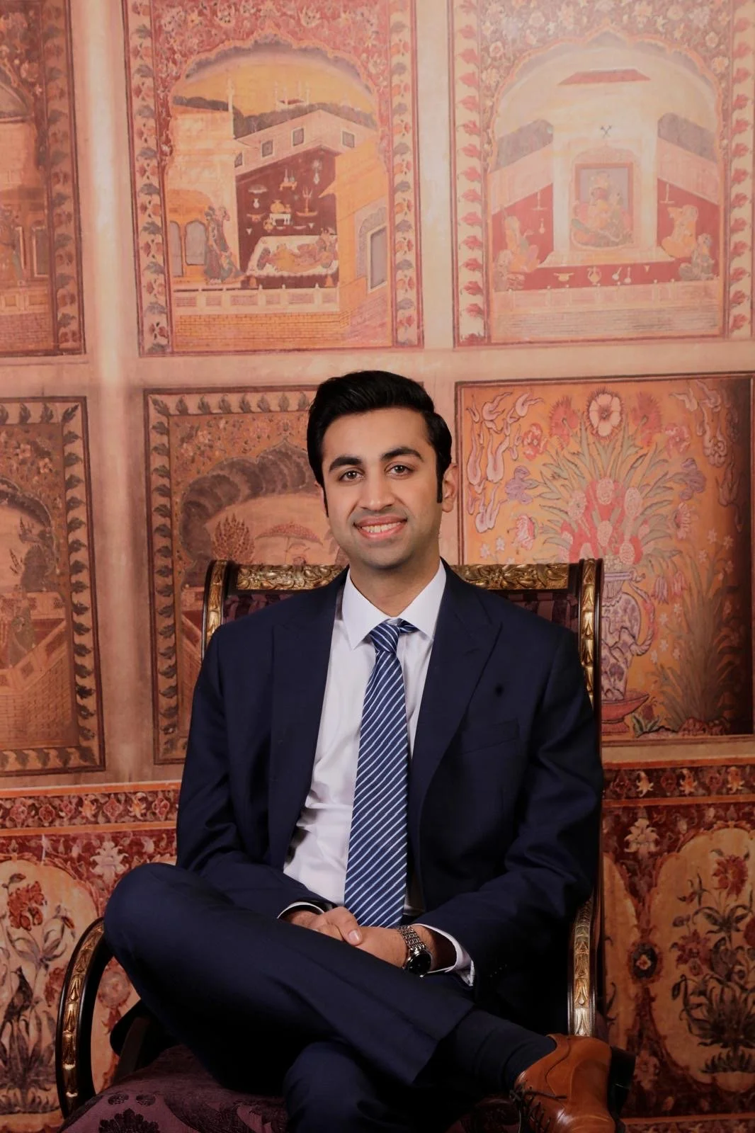 A man in a dark suit and striped tie sitting on a regal chair in front of an ornate, painted wall with floral and architectural designs.