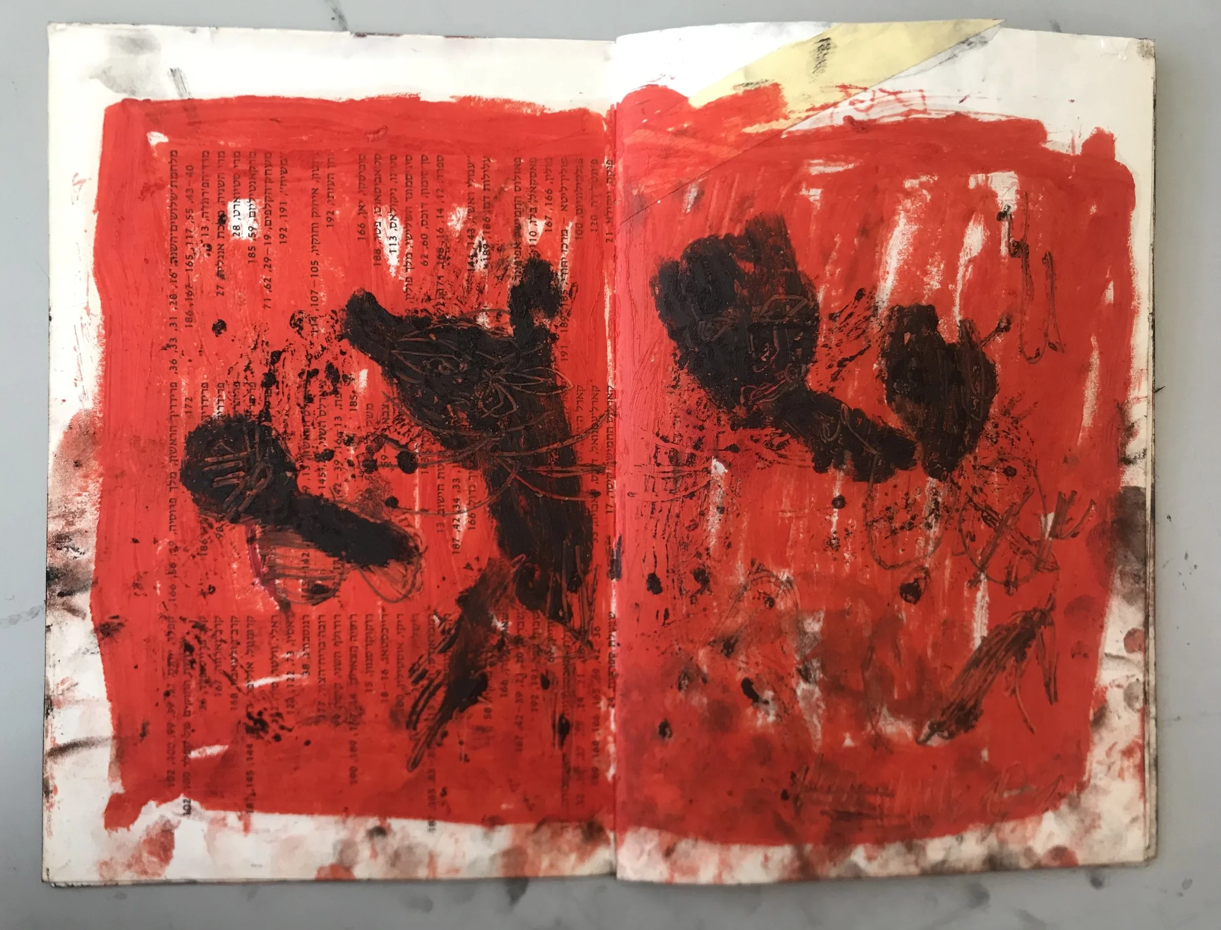 An open book with a red background on the pages, featuring black ink splatters, abstract shapes, and partially visible text in a language that appears to be Hebrew.