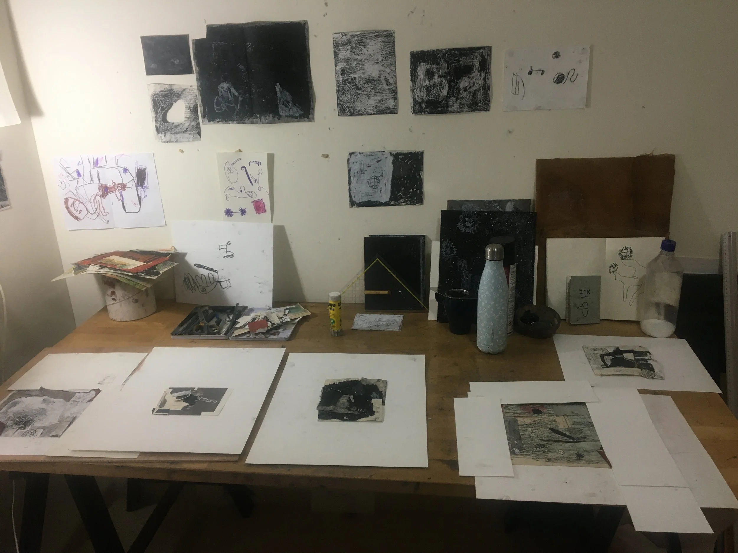 A workspace with black and white abstract artwork on the wall, small artwork and papers on the table, and various tools and items such as a water bottle, glue stick, and cans scattered around.