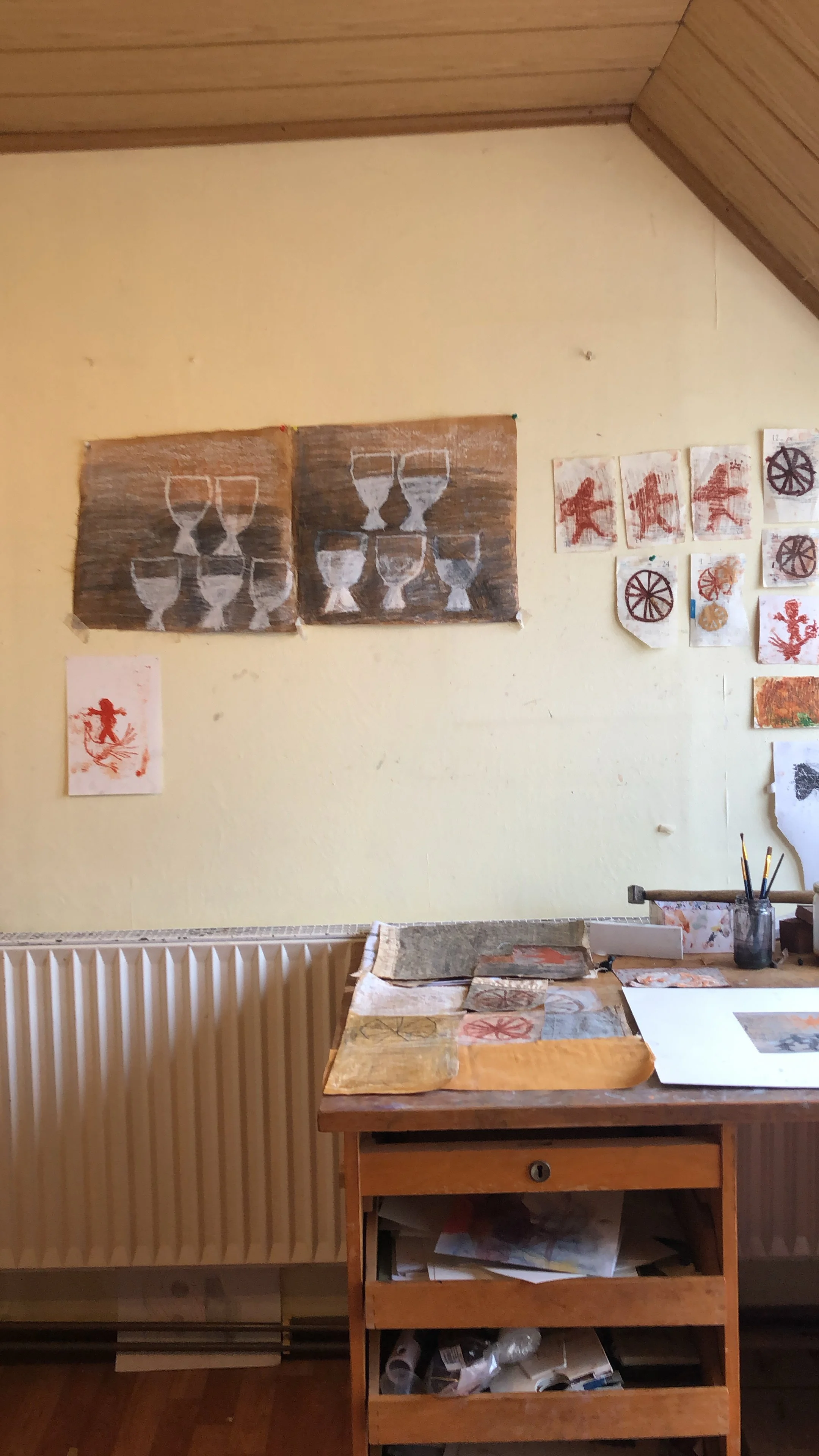 A room with a yellow wall and wooden ceiling, decorated with artwork featuring glasses and abstract designs. A wooden table holds various paper and art supplies, and a small rolling cart with multiple shelves contains art tools and materials.