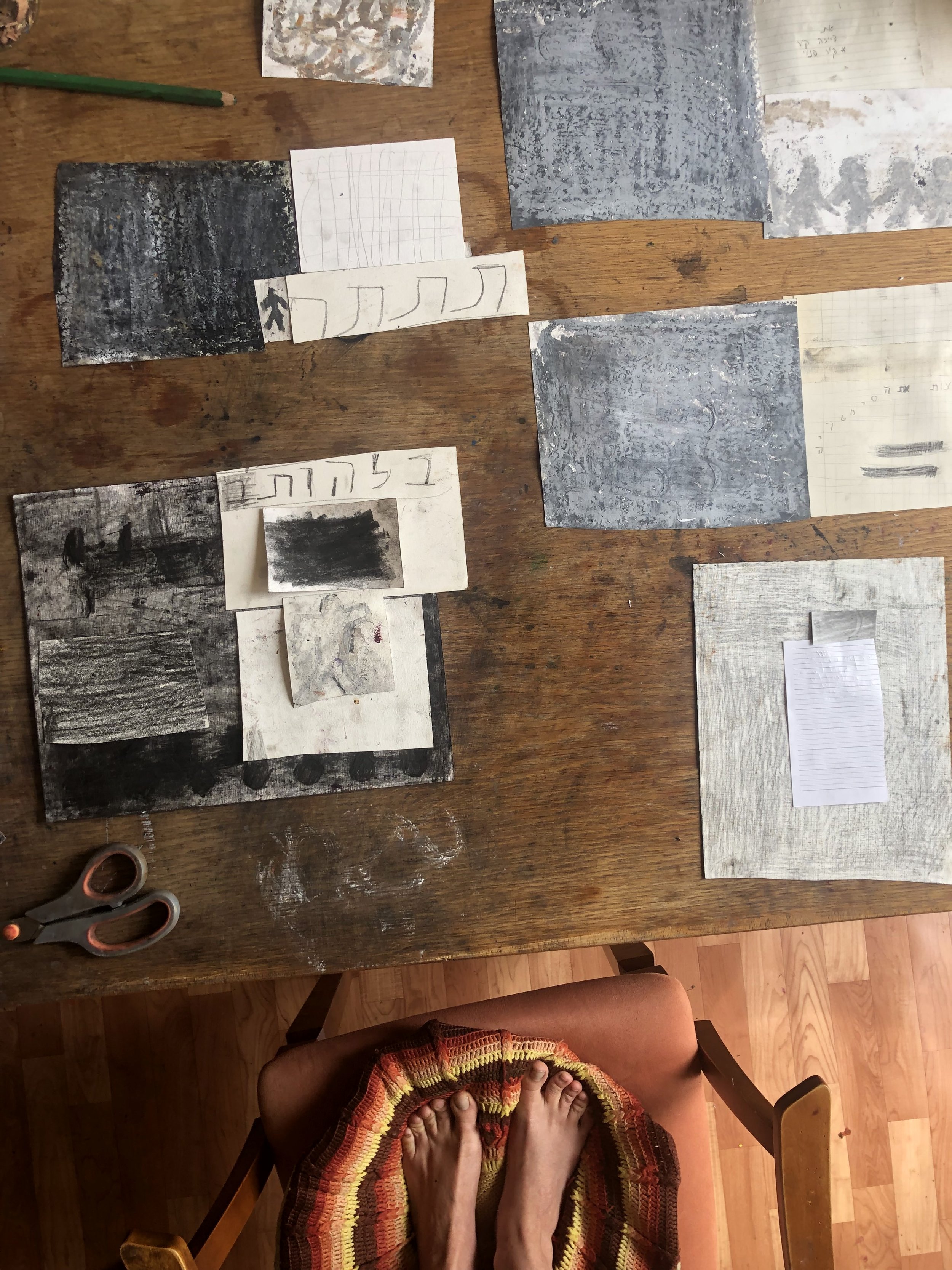 A person with bare feet sitting on a chair with a multicolored knitted blanket on their lap. A wooden table is in front of them with various pieces of paper and drawings, some with black and white textured images and Hebrew writing. There are scissor