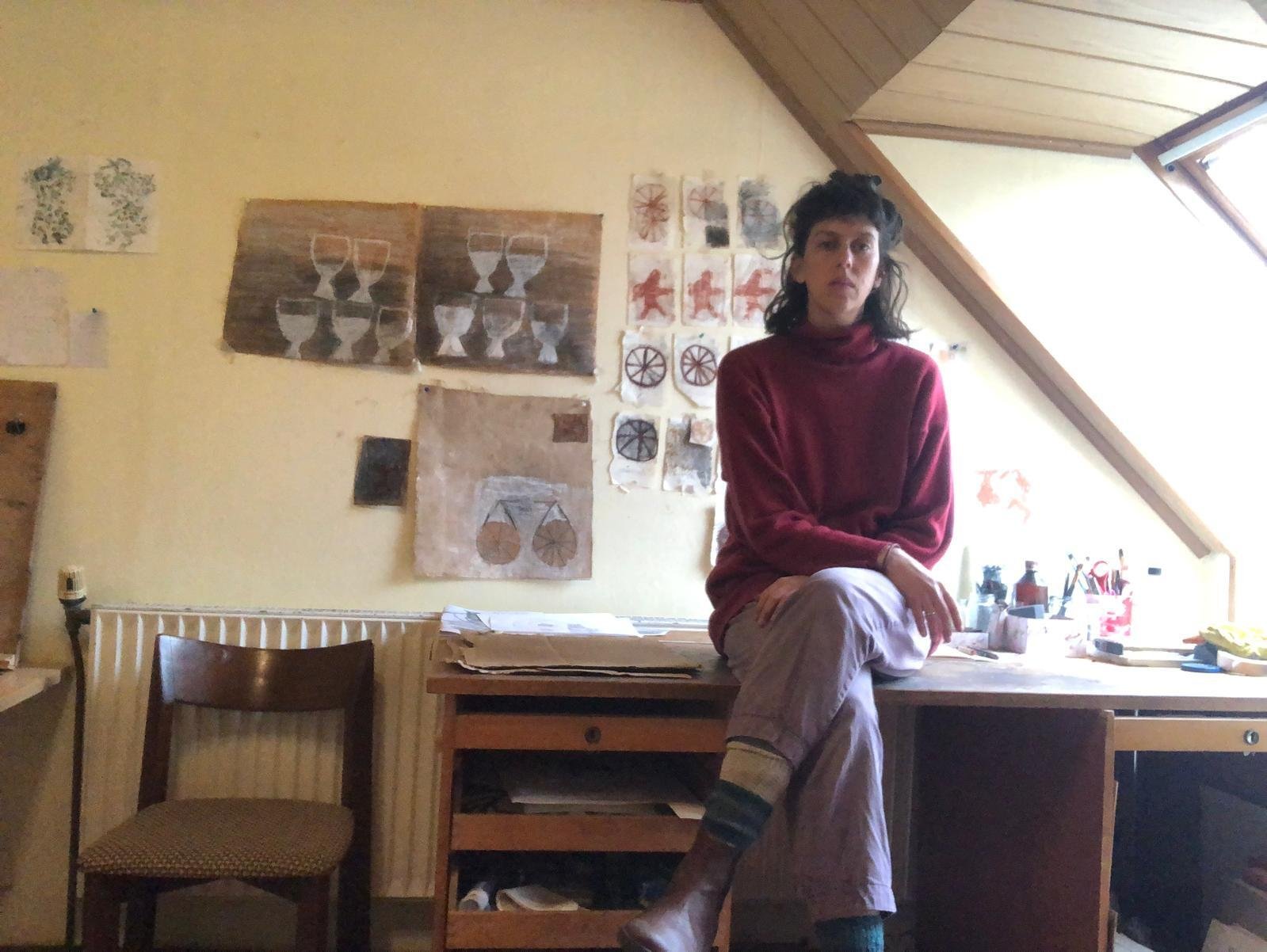 A woman in a red sweater and light-colored pants , artist yael shpinat , sitting on a wooden counter in a studio room with yellow walls decorated with artwork, natural light coming from a skylight window.