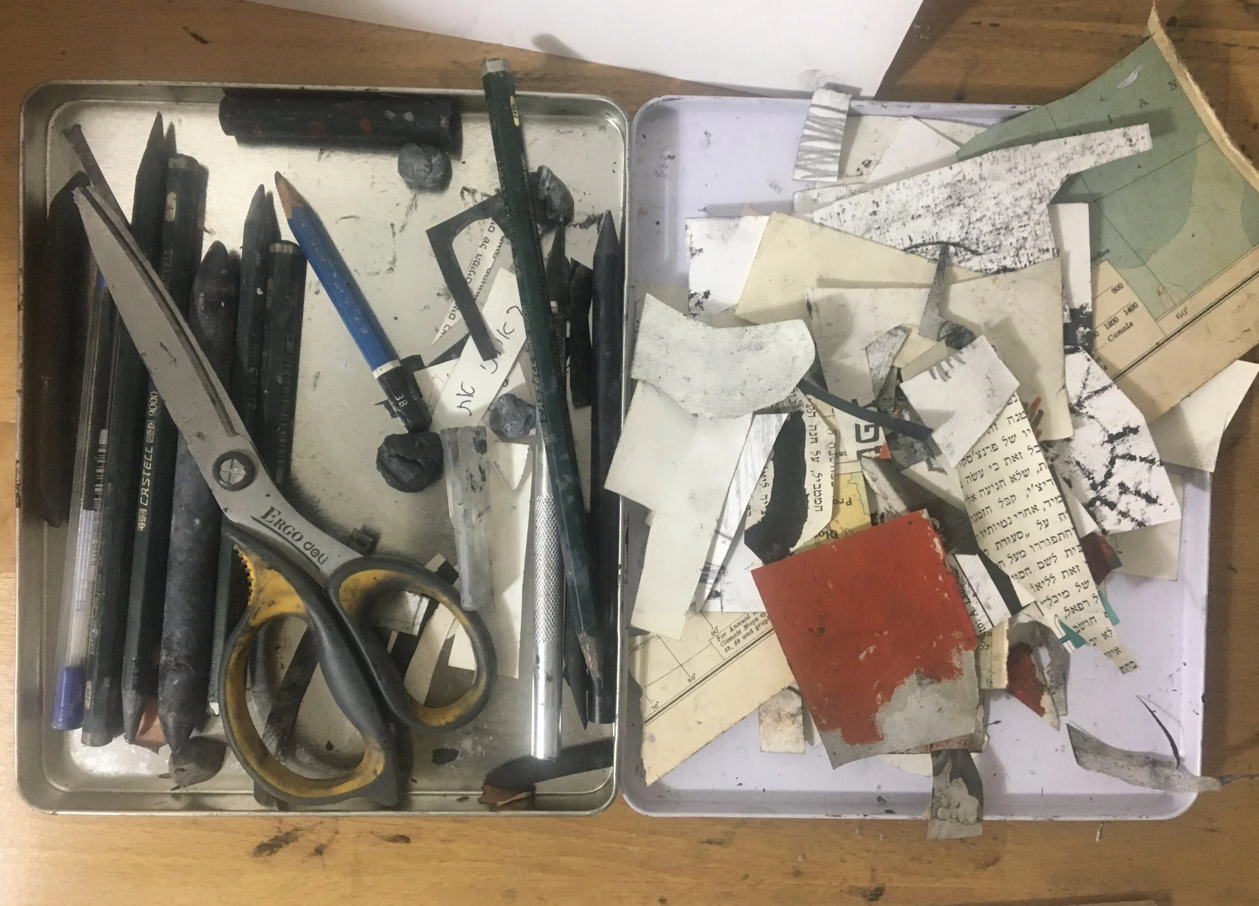 A metal tray containing various black markers, a green ruler, a pair of yellow-handled scissors, pebbles, and torn pieces of paper with handwritten and printed text. The tray is placed on a wooden surface.