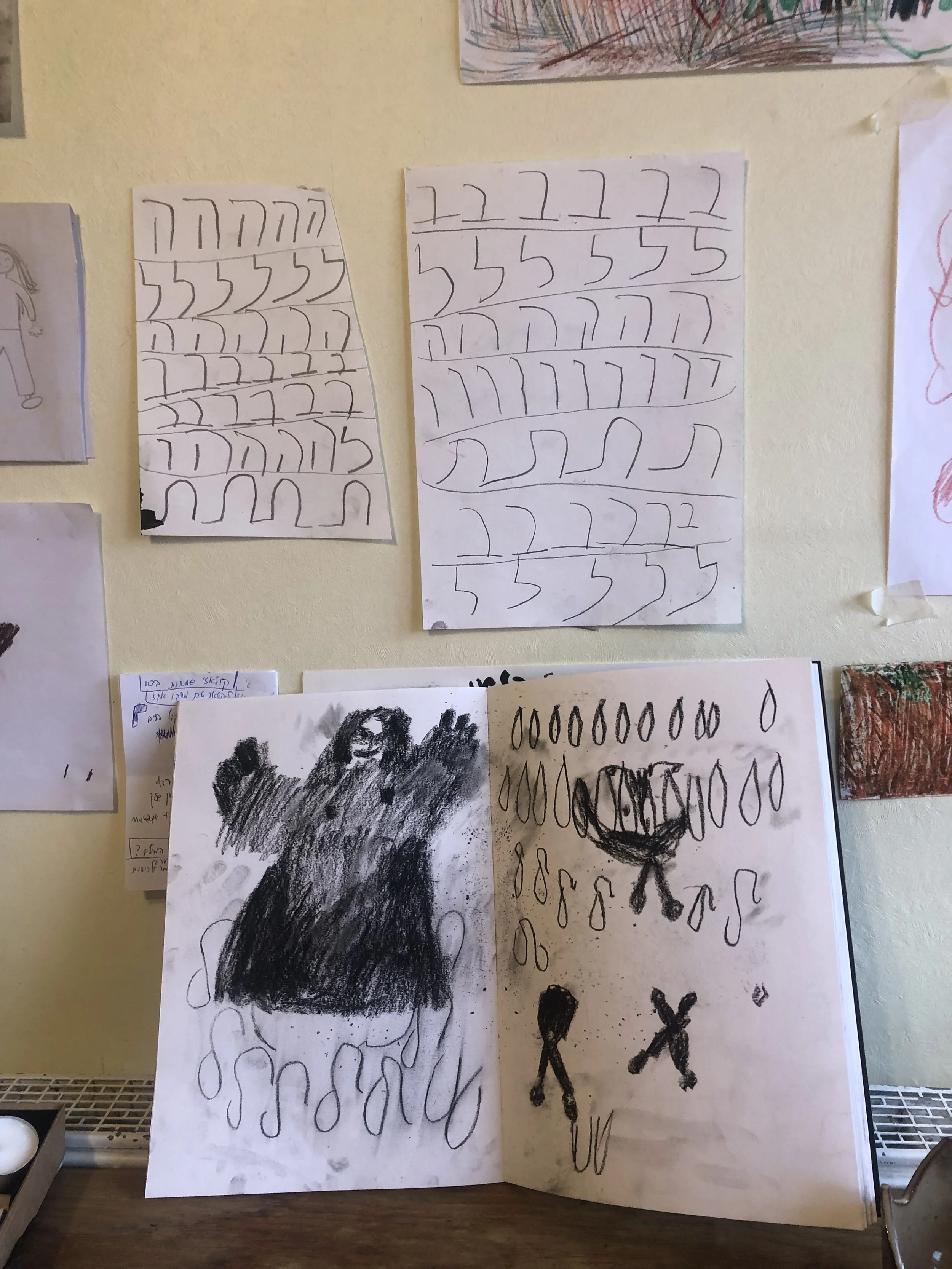 Children's artwork displayed on a wall, including drawings of faces, abstract shapes, and patterns made with markers and crayons.