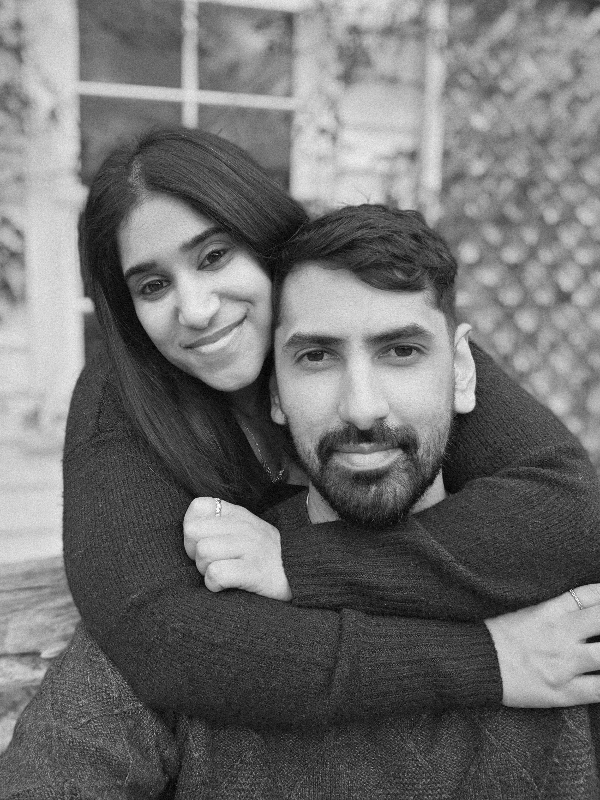 A black and white photo of a couple embracing outdoors, with the woman smiling and resting her head on the man's shoulder, and the man looking at the camera with a serious expression.