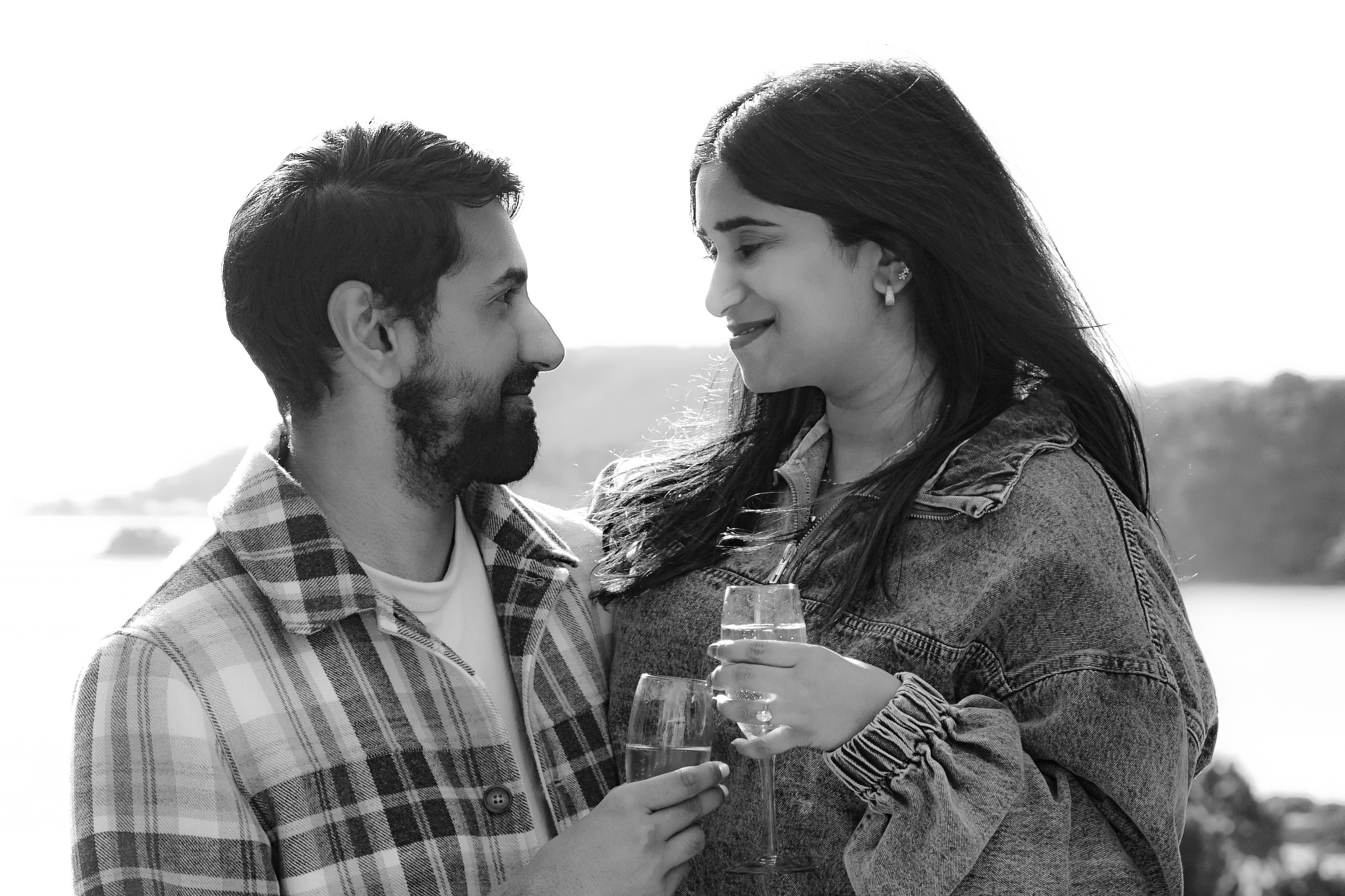 A black and white photo of a man and woman facing each other outdoors, smiling slightly, holding glasses of champagne in front of each other.