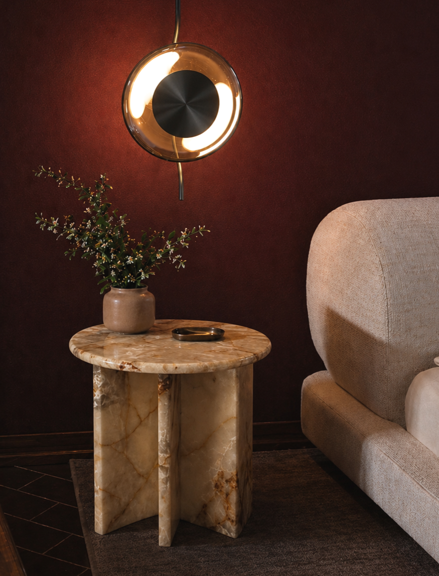 Living room corner with a round marble side table holding a brown vase with green foliage, a small black dish, a beige upholstered armchair, and a wall-mounted circular modern light fixture illuminating warm light.