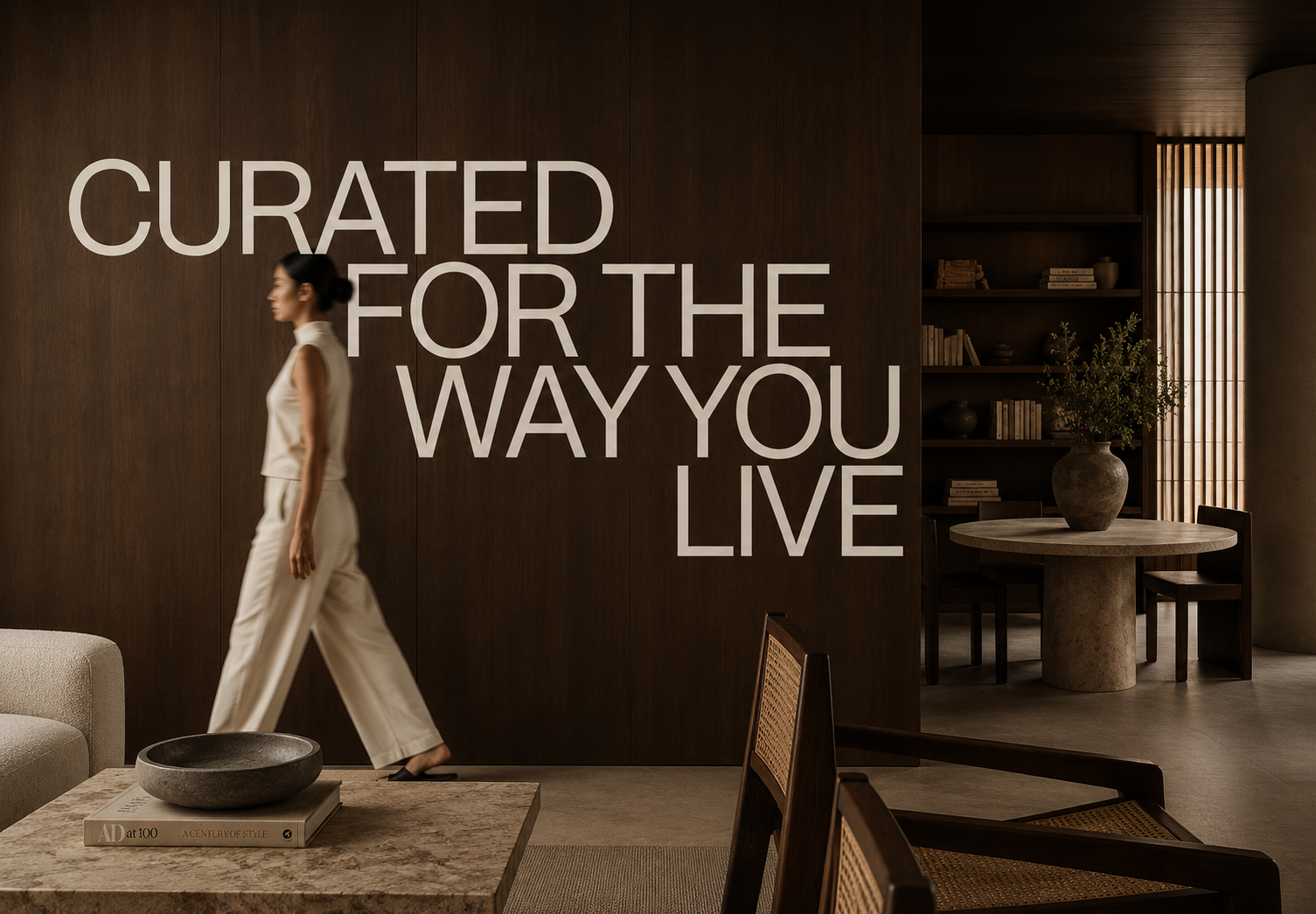 Interior of a modern, dimly lit space with a woman walking past a wall with large text that reads, 'Curated for the way you live.' There is a beige sofa, a marble table with books and a black bowl, and a round table with a large vase and plants in the background.