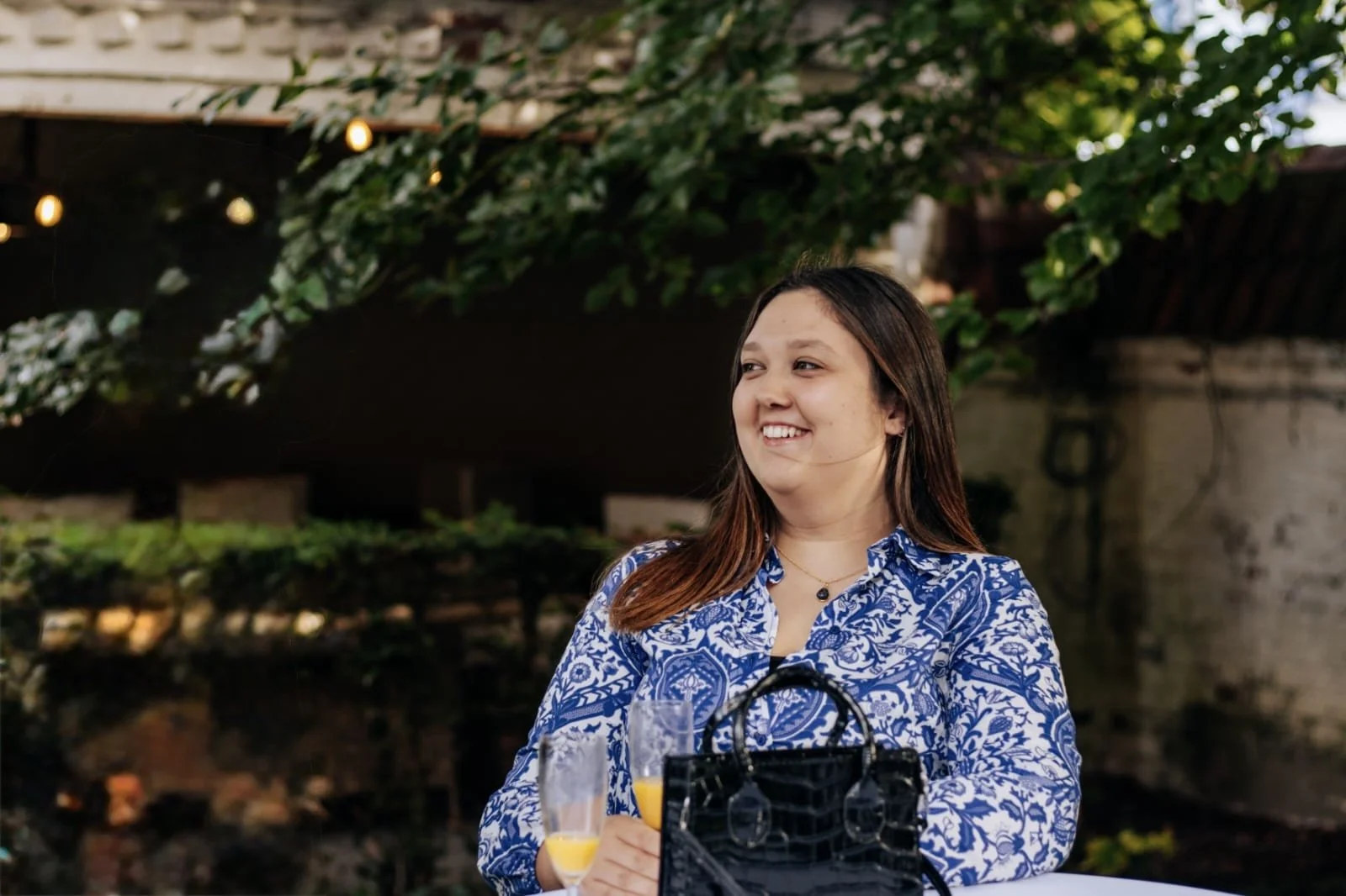 Vrouw met lang bruin haar, lachend, zit buiten, drinkt mimosas, draagt een blauwe en witte bloemenprint blouse, met een designer zwarte handtas op tafel, groene struiken en een muur op de achtergrond.