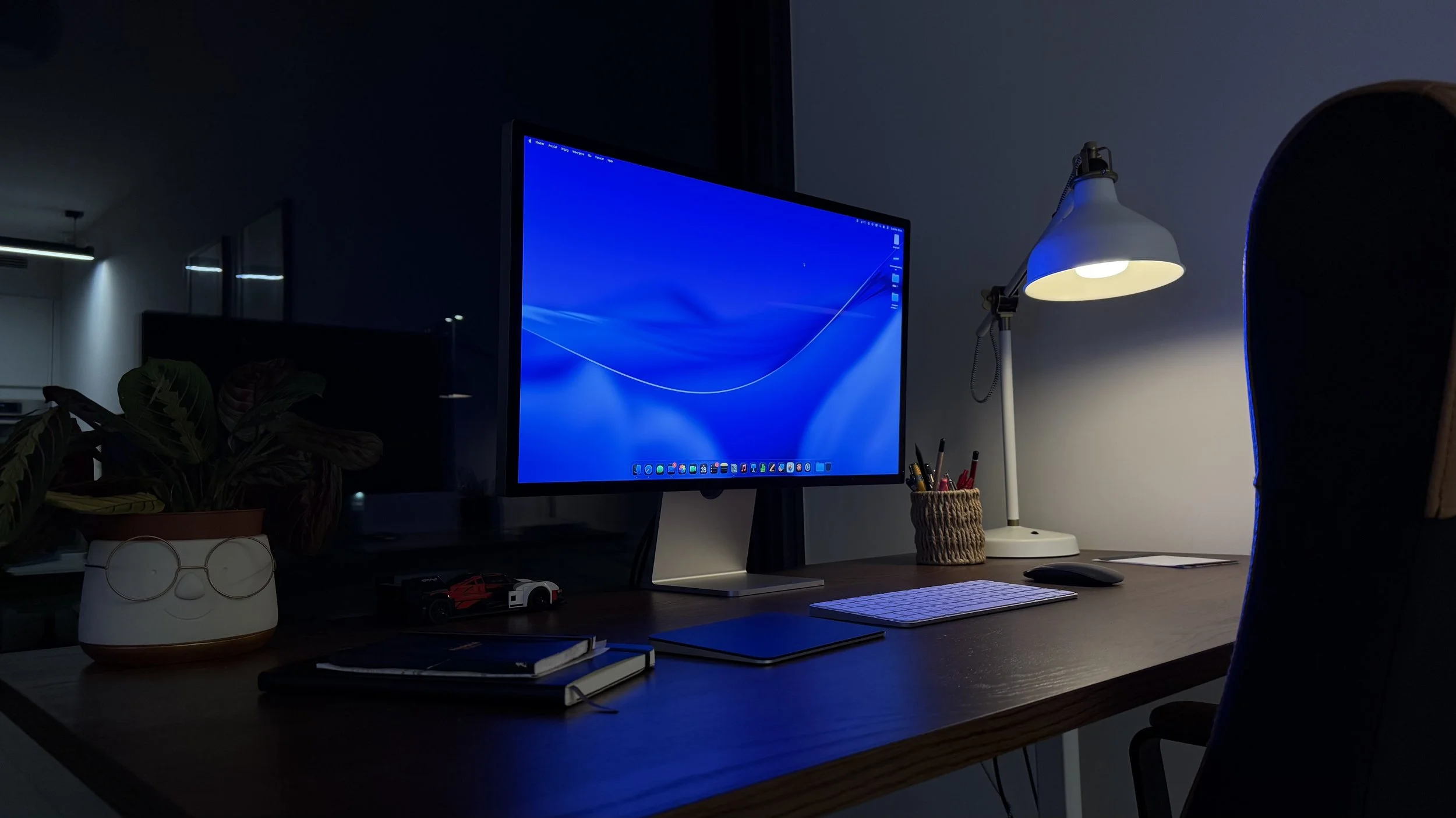 Een donkere thuissituatie met een bureau, een computer met een blauwe scherm, een bureaulamp met witte kap, een pot met potloden, een boek en een item met een gezicht met brillen
