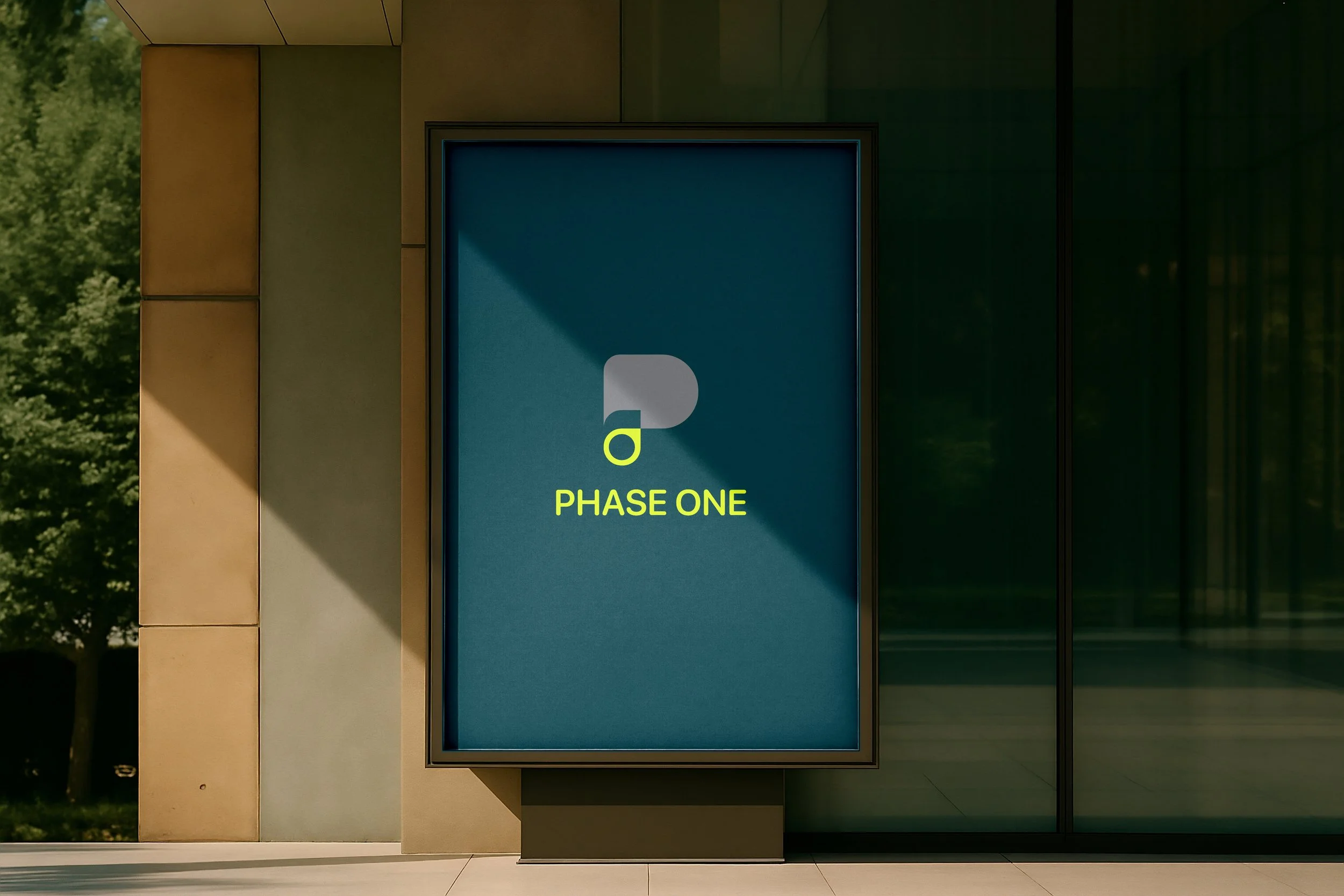 Digitale display met tekst 'PHASE ONE' en een logo, gelegen in een moderne gebouwfacade met glas en panelen, met groen buiten op de achtergrond.