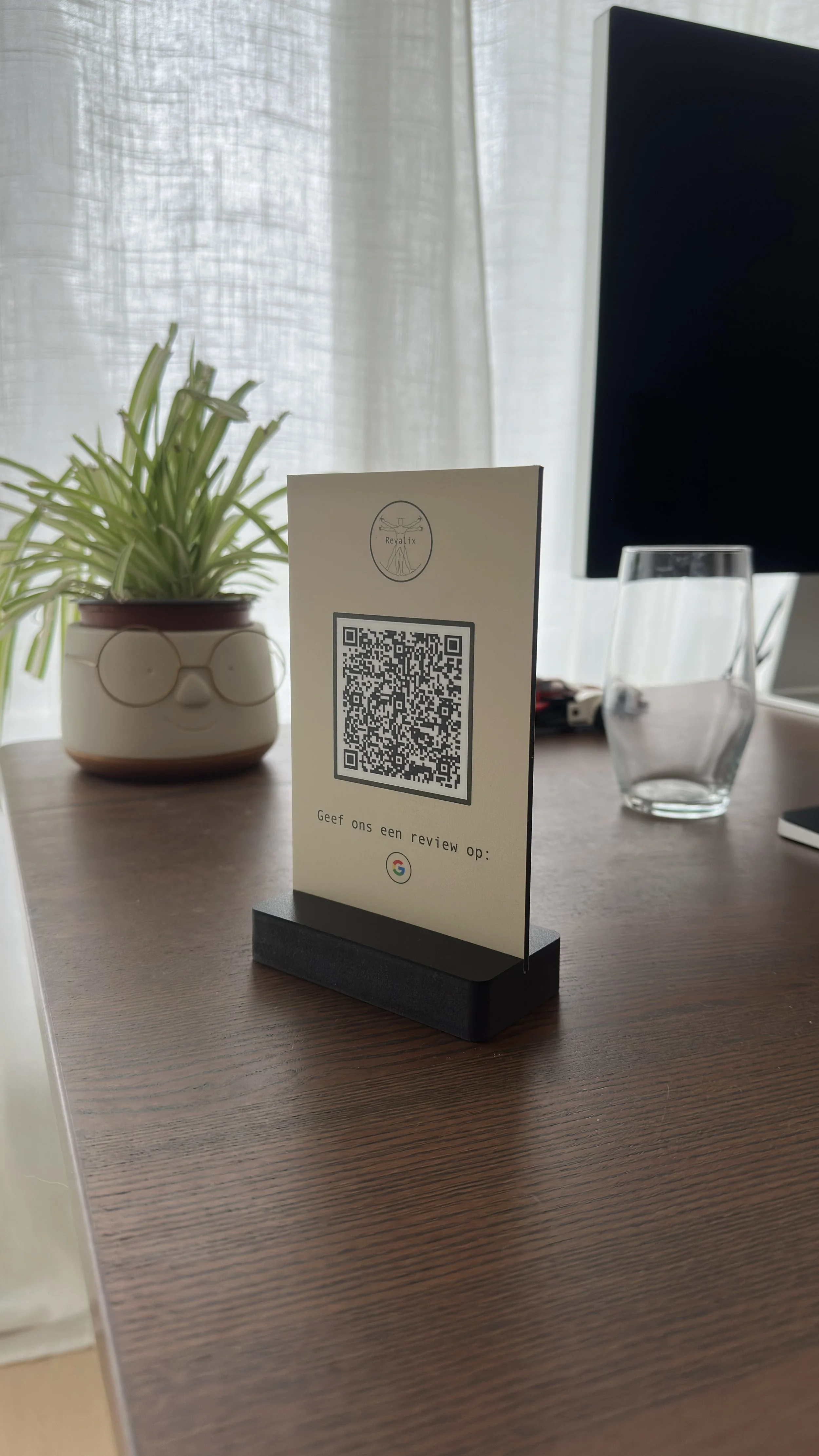Een tafel met een plantenpot met een groene plant, een informatiestand met QR-code, een lege waterglas en een monitor. Achtergrond met lichte gordijnen.