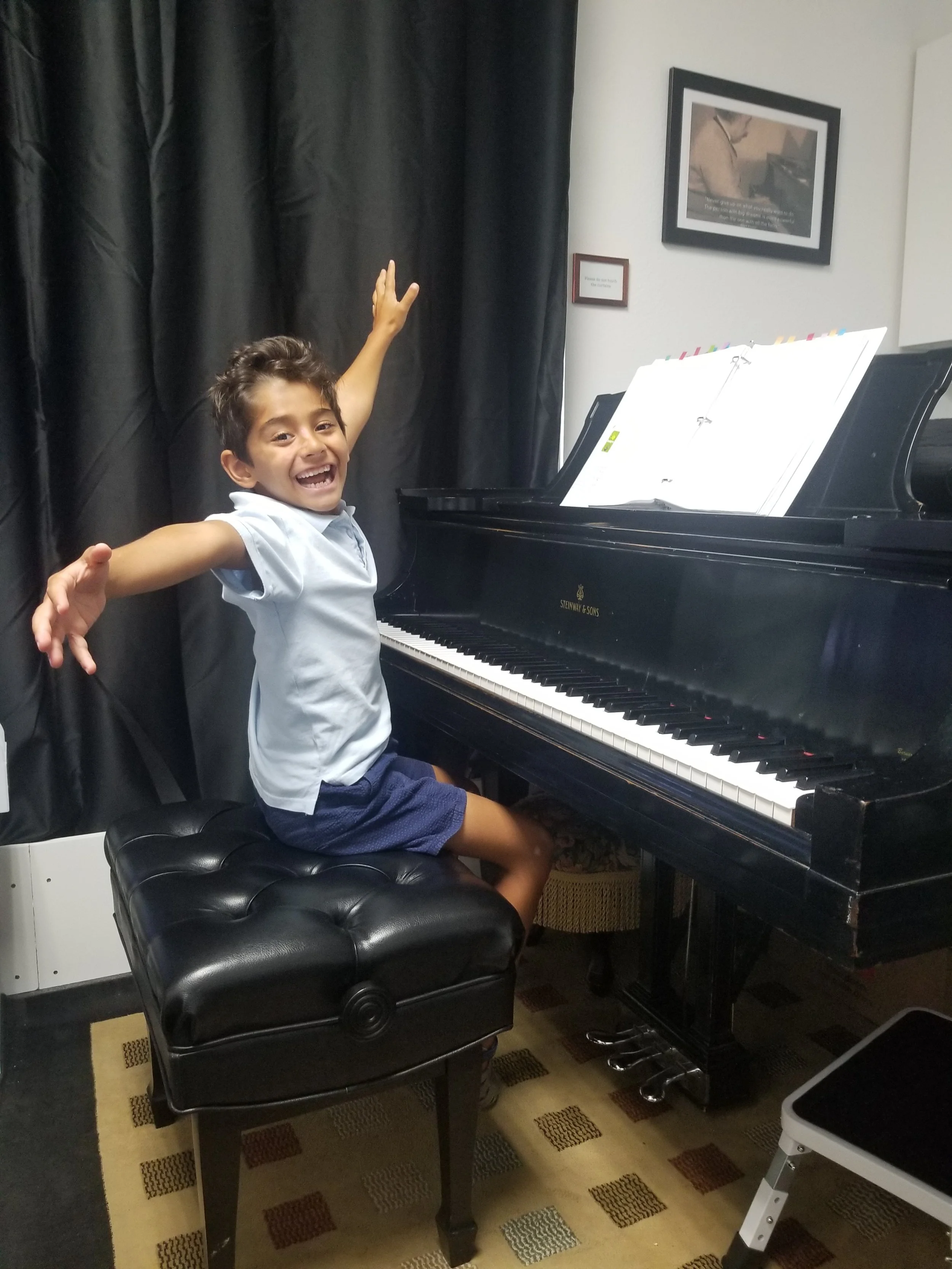A young boy with dark hair smiling and raising his arms, sitting on a black piano bench next to a black grand piano with sheet music. The room has a white wall with framed pictures and a dark curtain to the left.