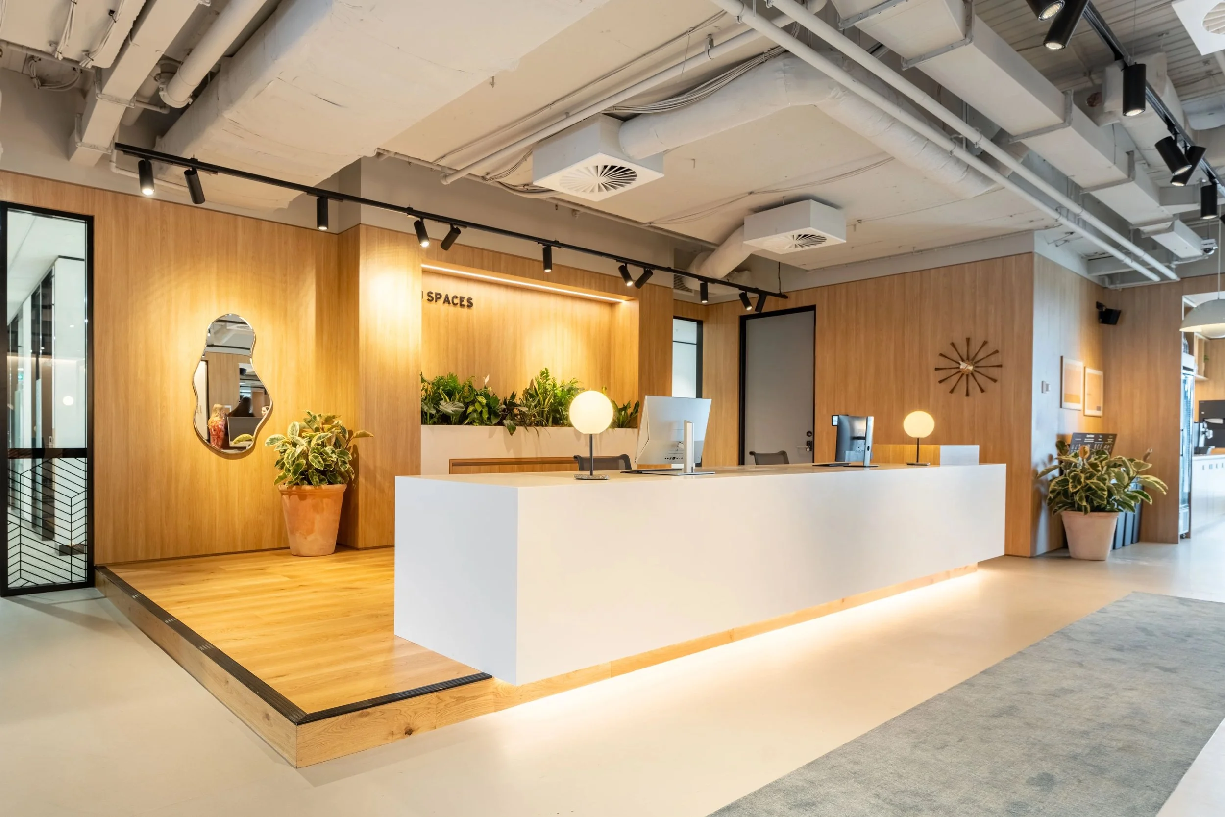 Modern office reception desk with a white counter, potted plants, wall-mounted mirror, and wood-paneled walls in a contemporary setting with track lighting and ceiling ducts.