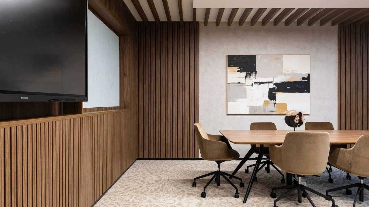Modern conference room with wooden panel walls, a large abstract painting, six beige chairs around a wooden table, a black decorative sculpture, a patterned carpet, and a flat-screen TV on the wall.