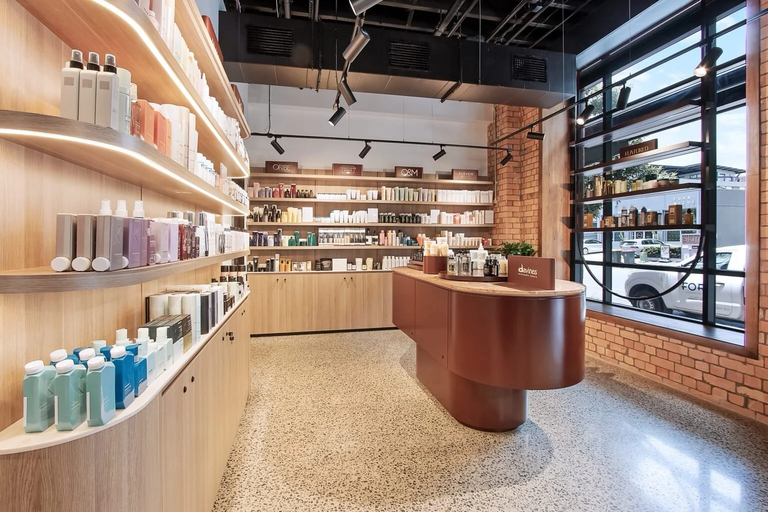 Interior of a modern retail store with wooden shelves stocked with beauty products, a curved wooden checkout counter, large window with brick wall, and track lighting.