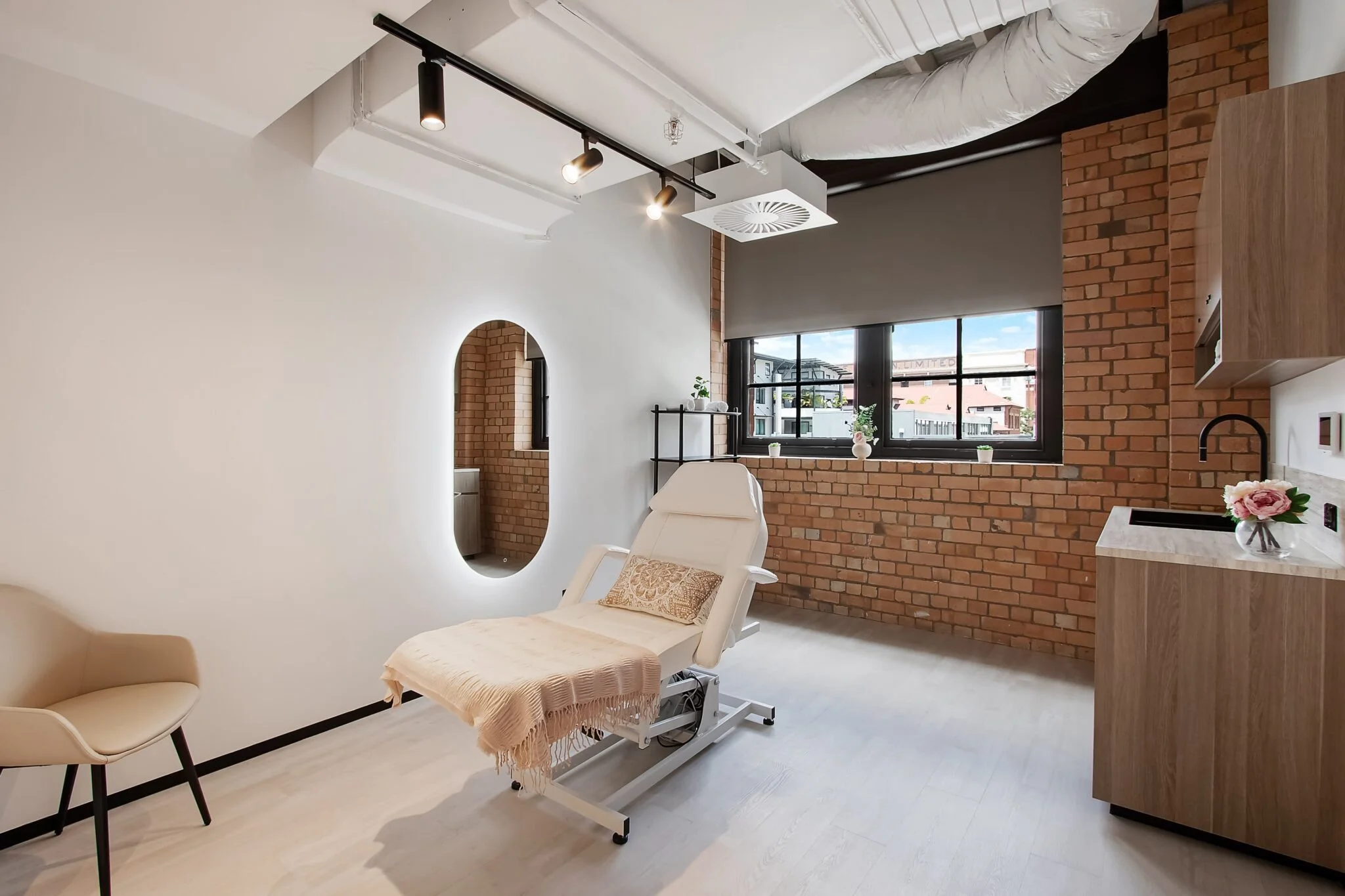 A modern room with exposed brick walls, large window, and medical equipment including a patient bed with a beige blanket and an angel wing pillow, a beige chair, a small table with flowers, and wall-mounted shelves.