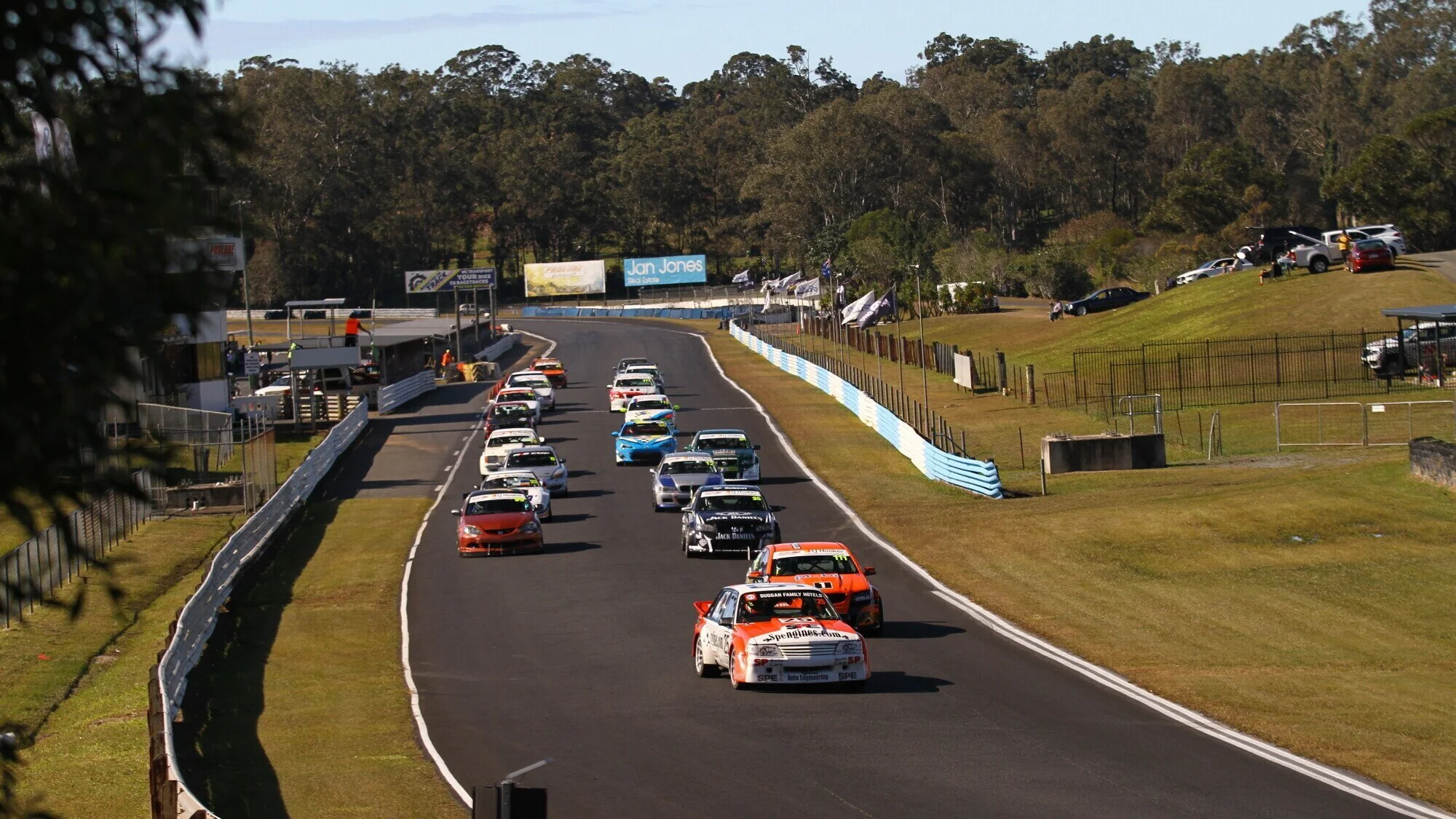 Race cars on a race track during a motorsport event, with spectators' cars parked on a hill in the background and trees beyond.