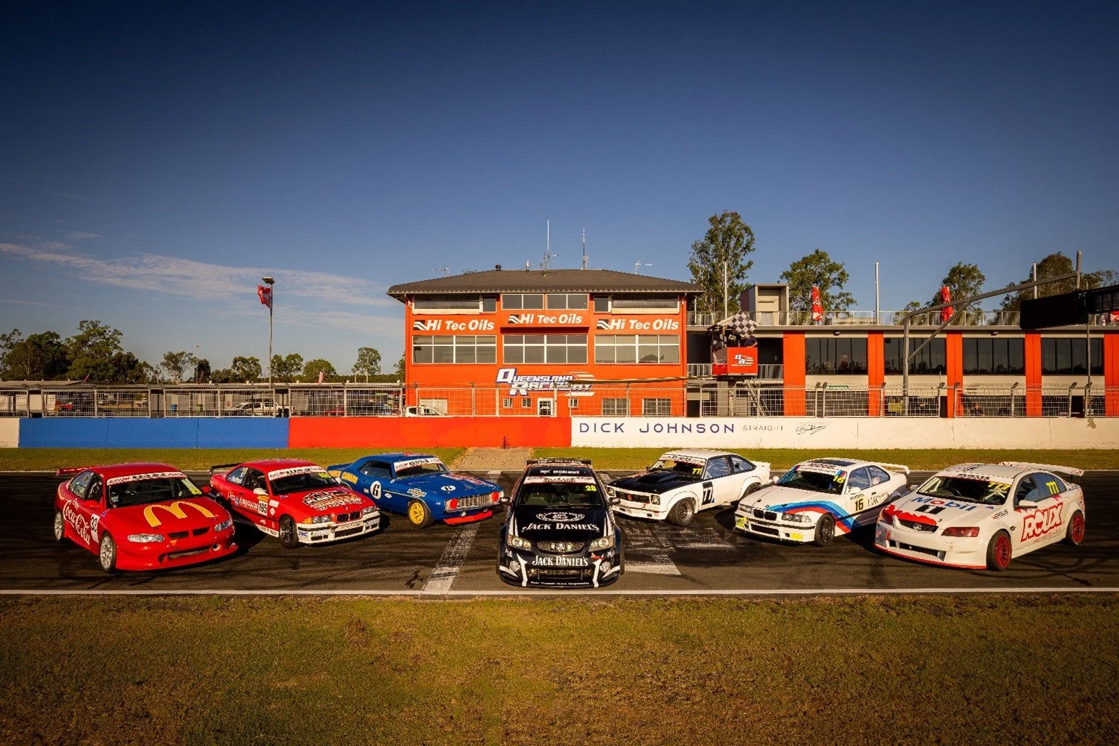 Group of race cars parked on a race track with a building in the background that has Hi Tec Oils branding, flags, and trees in the distance.