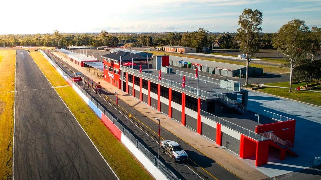 An aerial view of a race track with a red and black building, bleachers, and a parking lot in the background.
