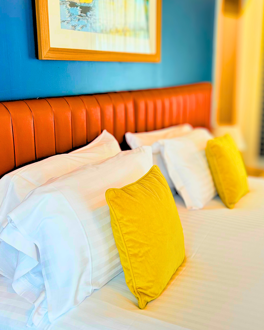 A close-up of a neatly made bed with white pillows, yellow accent pillows, a red leather headboard, a blue wall, and framed artwork above the bed.