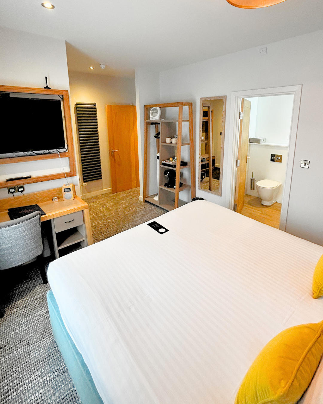 A hotel room with a large bed, a flat-screen TV on the wall, a desk with a chair, a small kitchenette with shelves, and a bathroom visible through an open door. The room has wooden furniture and a neutral color scheme.