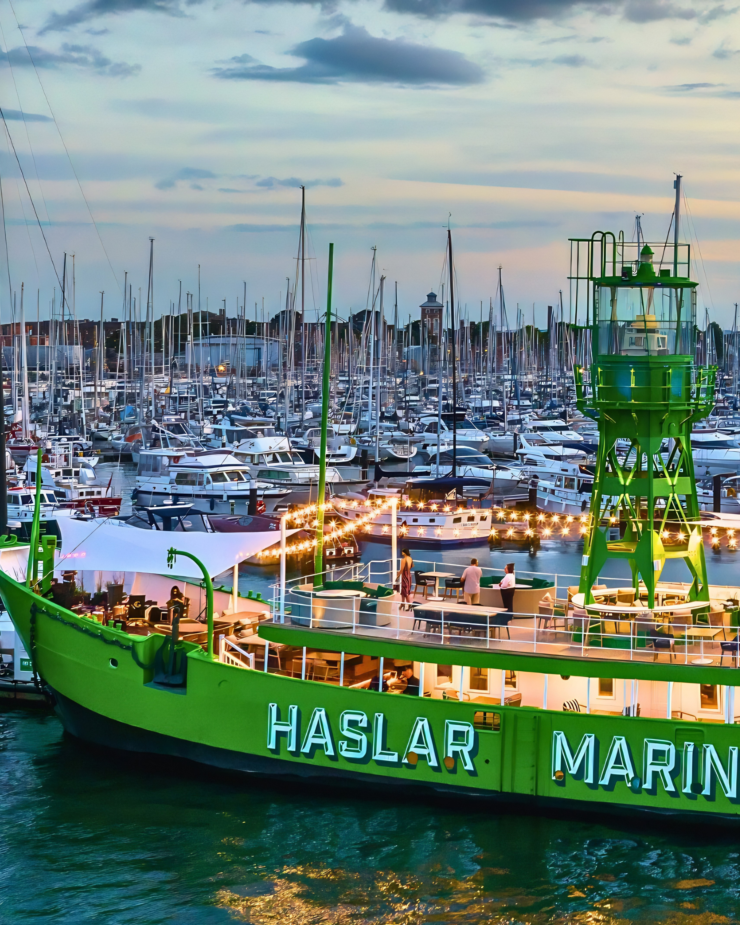 A large green boat named HASLAR MARIN docked at a marina filled with numerous sailboats and yachts, with a partly cloudy sky overhead and some buildings in the background.