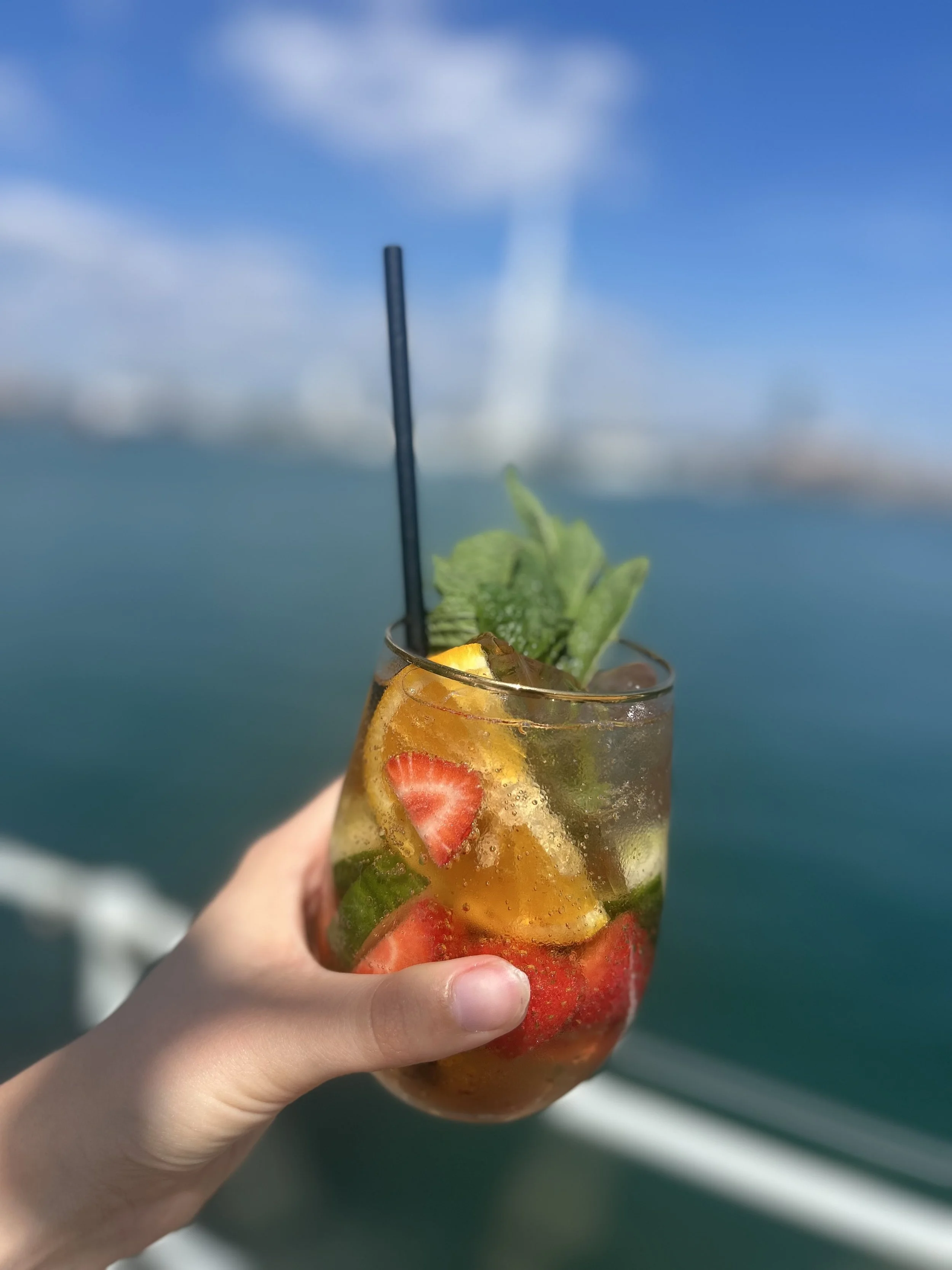 Hand holding a glass of colorful fruit cocktail with strawberries, mint, and orange slices, with a blurred ocean and sky background.