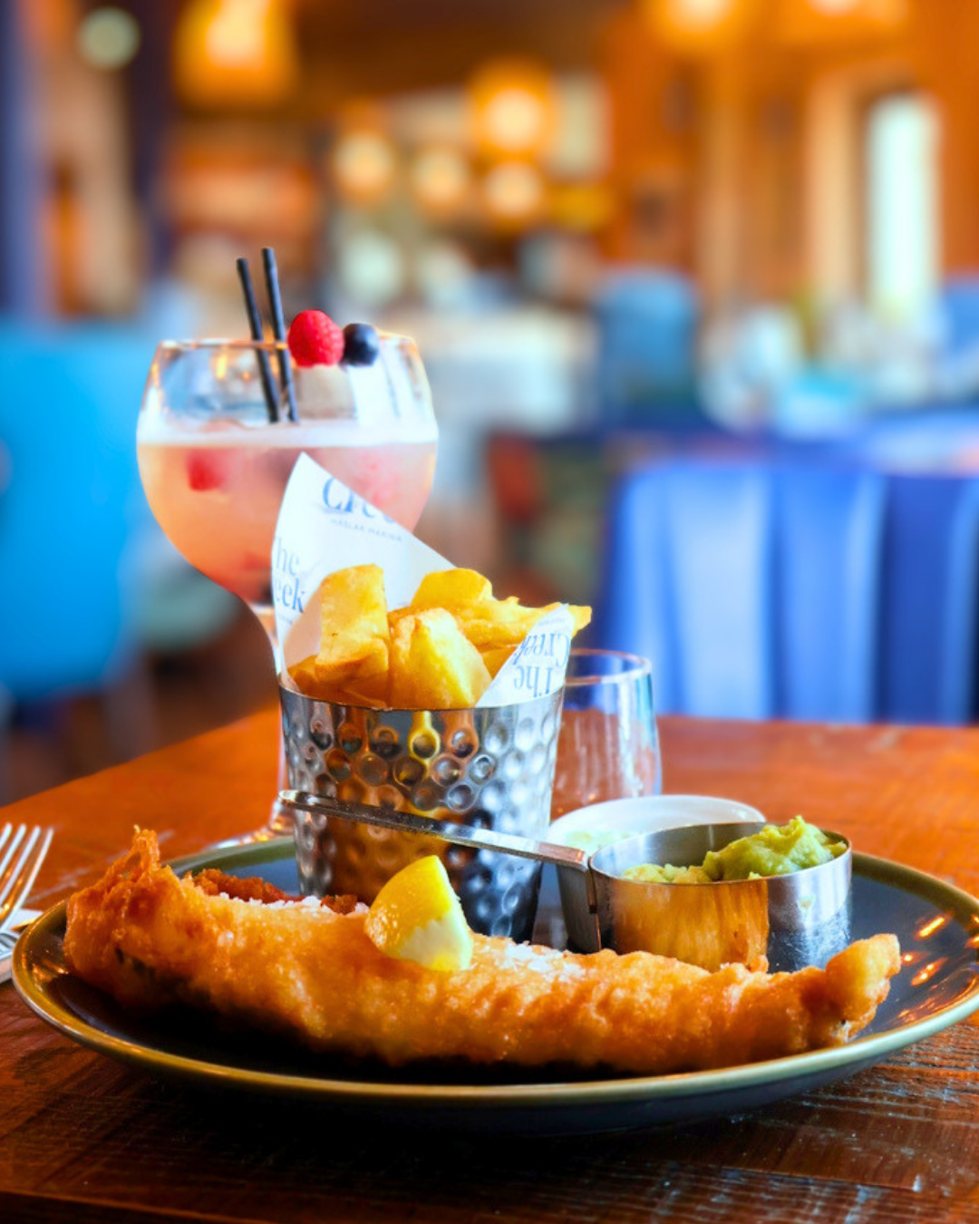 Plate with fried fish, lemon wedge, side of mashed peas, a cup of fries, and pink cocktail with berries and black straws on a wooden table in a restaurant.